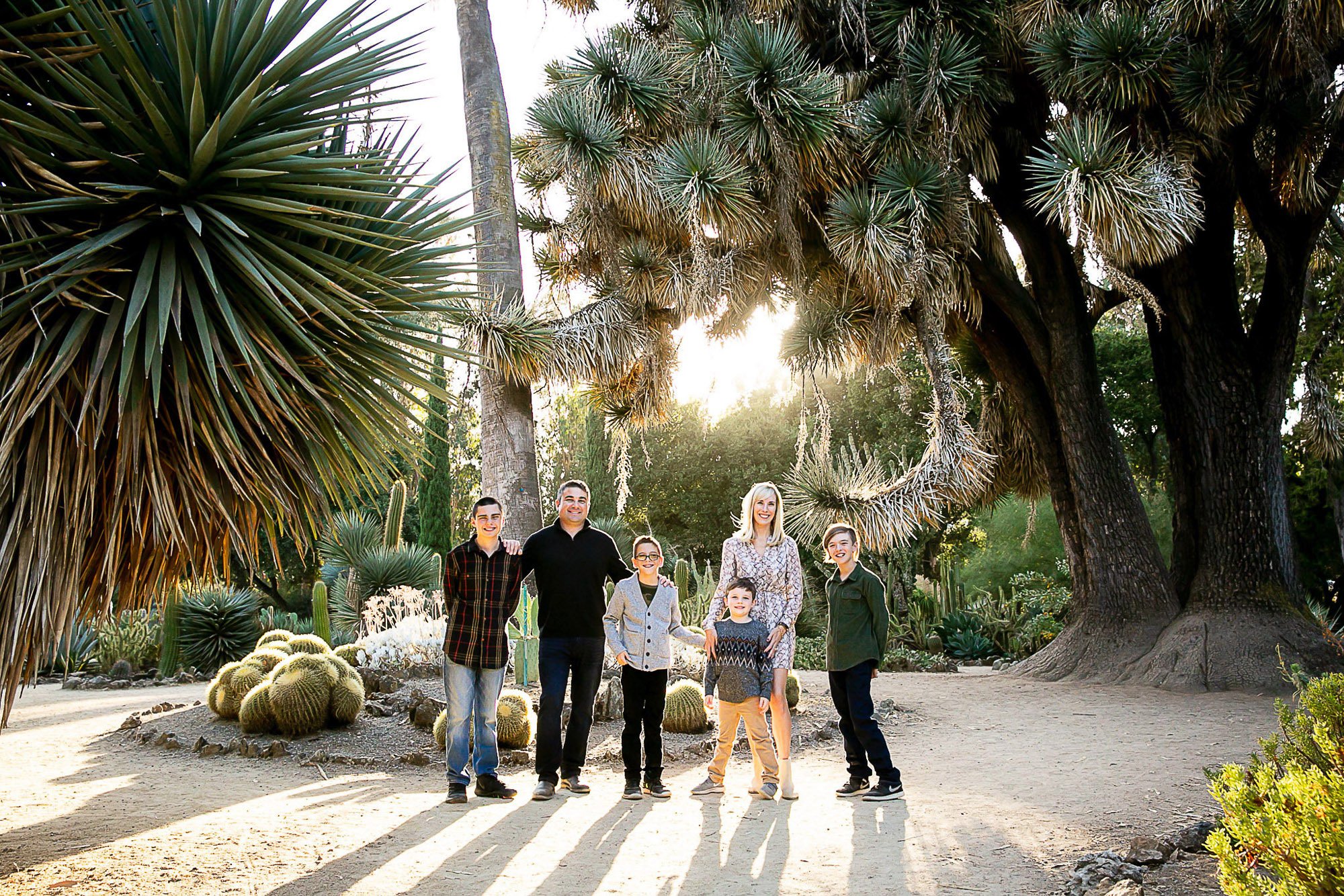 A family of eight standing in a desert botanical garden with large succulents and cacti, and tall trees, during sunset.