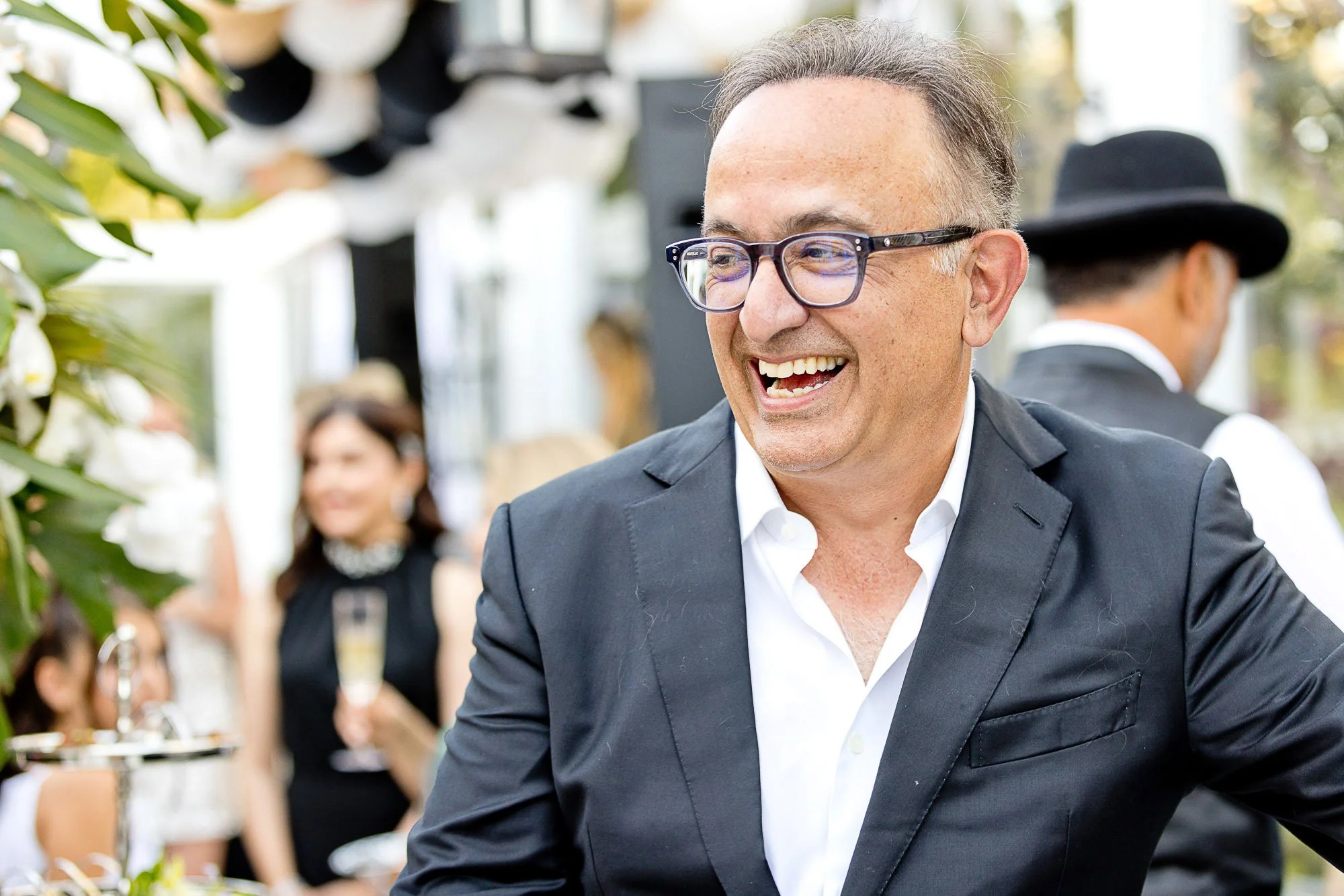 A man with glasses smiling and wearing a dark suit at a social gathering, with a woman in a black hat and dress in the background.