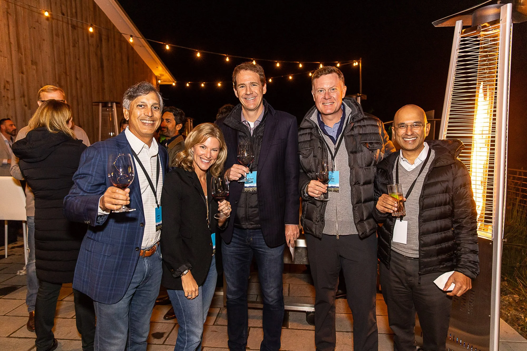 Group of seven people at an outdoor evening event, some holding wine glasses, standing near a patio heater with string lights overhead.