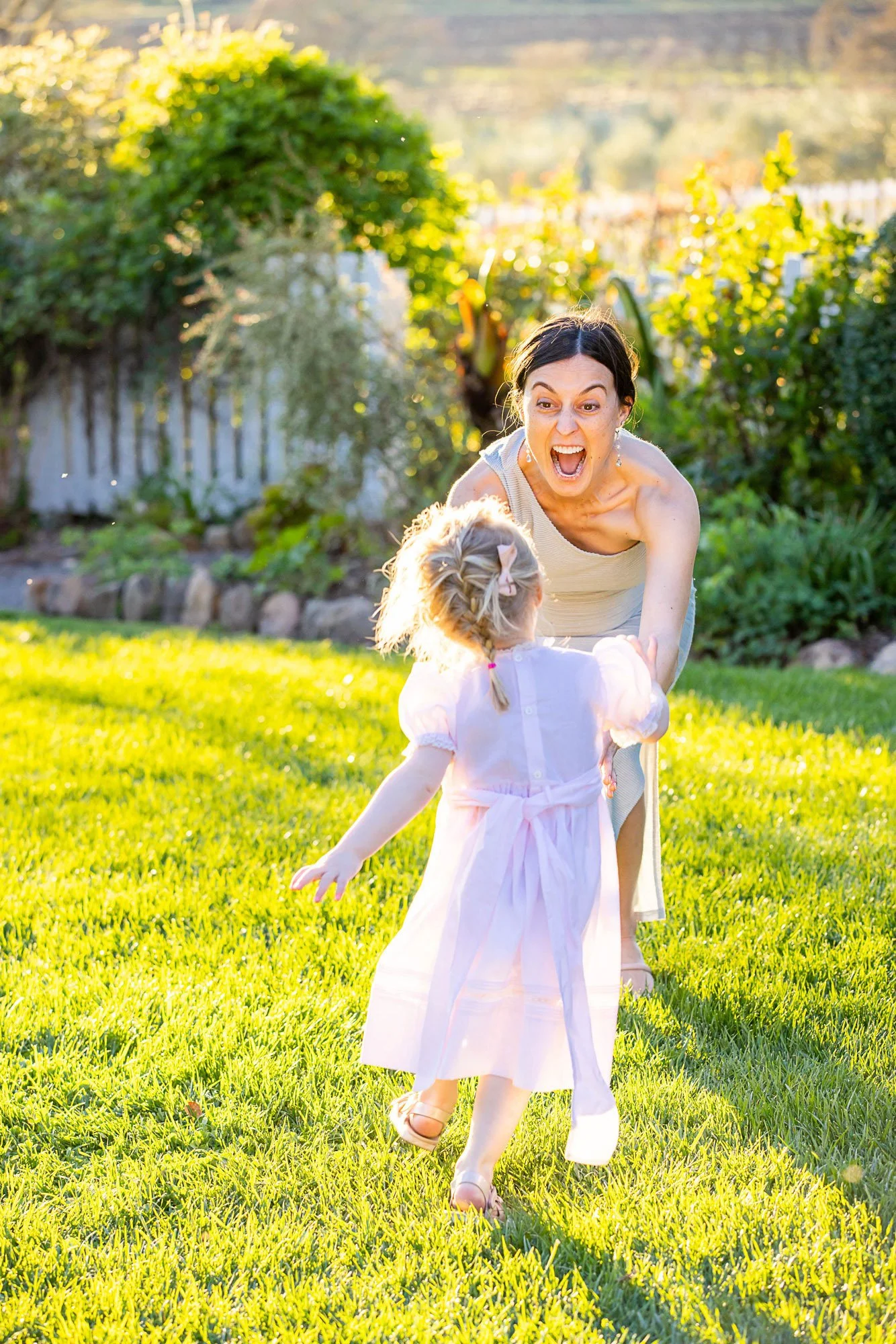 A woman and a young girl are playing together on a grassy backyard on a sunny day. The woman appears to be excited and is reaching towards the girl, who is facing away from the camera. The background shows trees and a white picket fence.