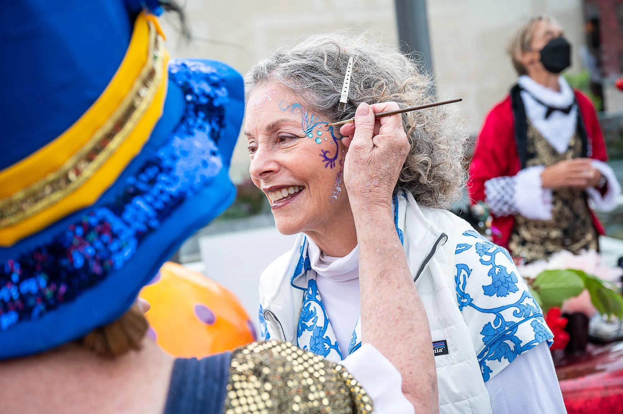 An elderly woman with curly gray hair and a broad smile painting a blue and purple design on the face of a person wearing a colorful hat. Other people, some wearing masks, are visible in the background at an outdoor event.