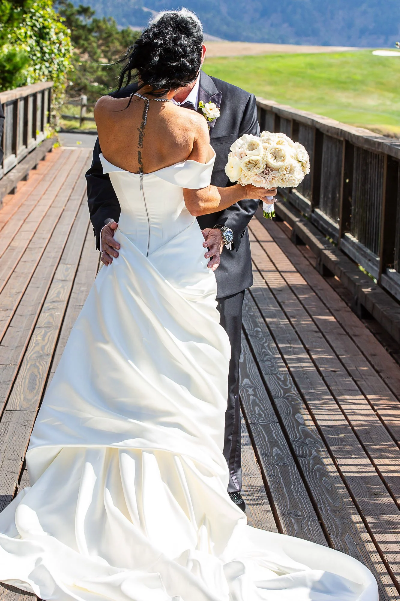 A bride and groom at the Beach and Tennis Club in Pebble Beach, CA on a wooden outdoor walkway, with the groom dressed in a dark suit and the bride in a white wedding gown holding a bouquet of white roses.