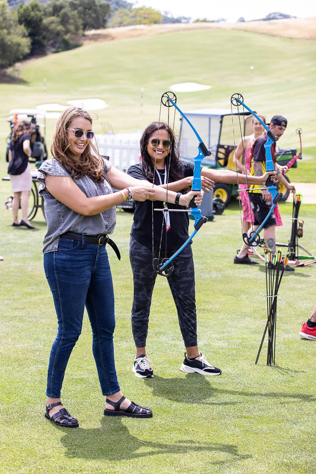 Two women smiling and holding archery bows, standing on a grassy field with archery equipment and other people in the background.
