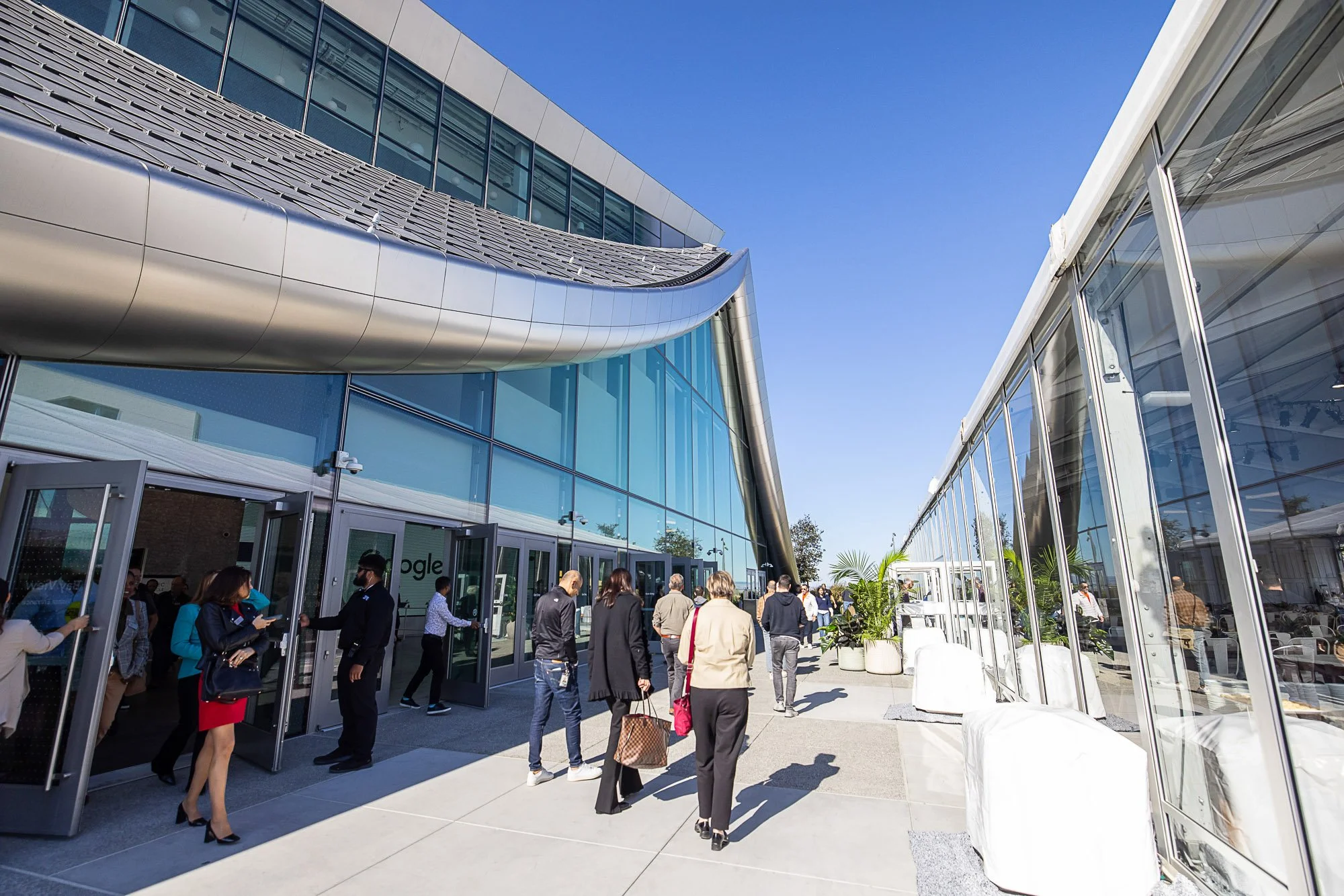 People entering and gathering outside a modern building with glass walls and a metallic, wave-shaped roof under a clear blue sky.