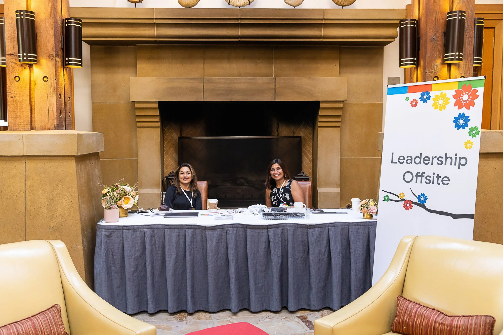 Two women sit behind a table at a leadership offsite event in a room with a fireplace. The table is covered with papers and has floral arrangements on it. To the right, there is a colorful banner that reads 'Leadership Offsite' with a floral design.