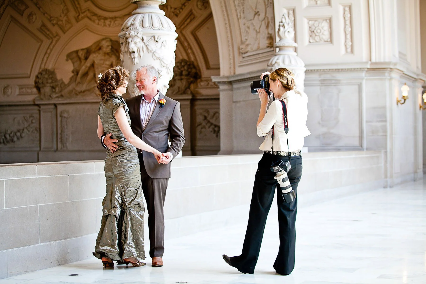 A woman and a man are posing for a photo in an elegant, classical building with ornate sculptures and architectural details. The woman is wearing a shiny, textured gown, and the man is in a suit with a boutonnière. Arrowood is taking their picture.