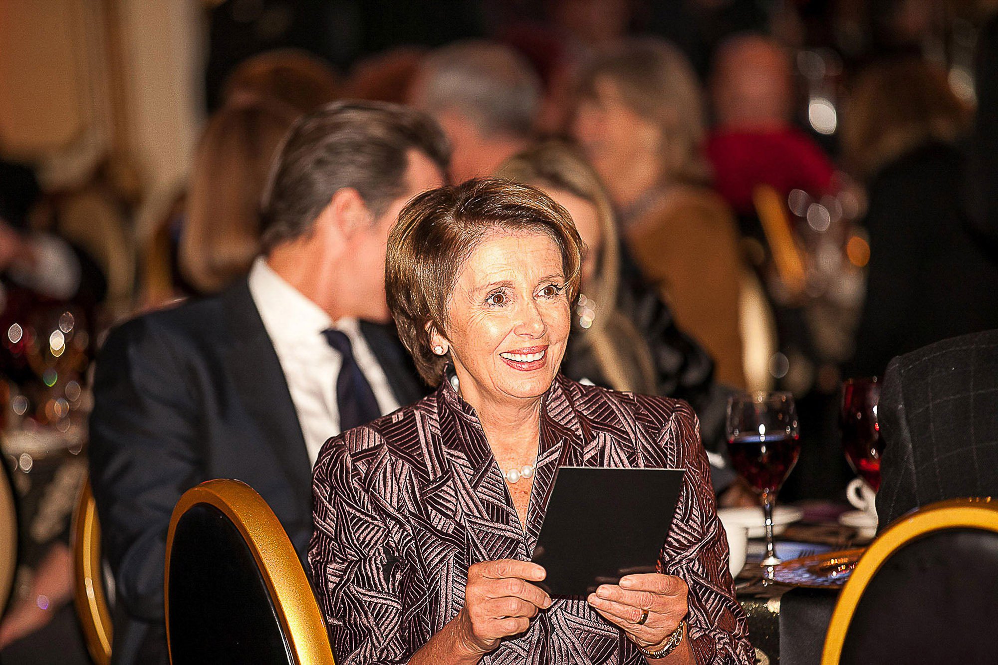 Nancy Pelosi, wearing a patterned maroon blazer, smiling while holding a glass of red wine, seated at a table with a man in a dark suit and tie, in a dimly lit banquet or conference setting with other seated guests in the background.