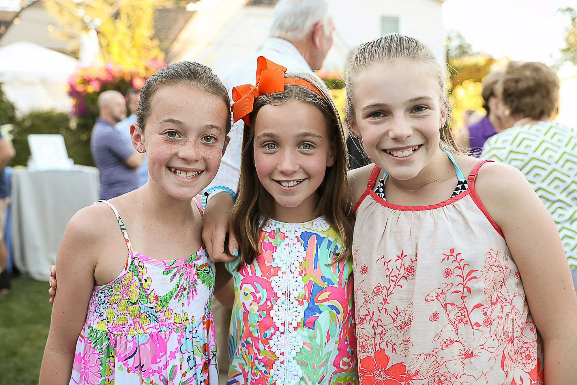 Three young girls smiling and posing for a photo at an outdoor gathering, with other people and decorations in the background.
