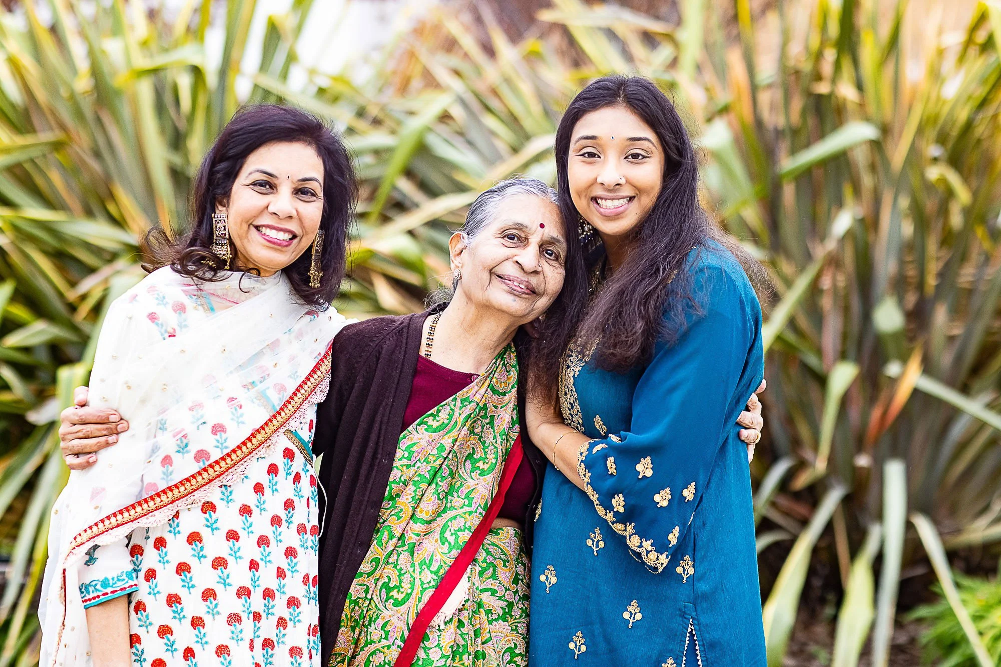 Three women standing together outdoors in front of tall green plants, smiling, wearing traditional Indian clothing.