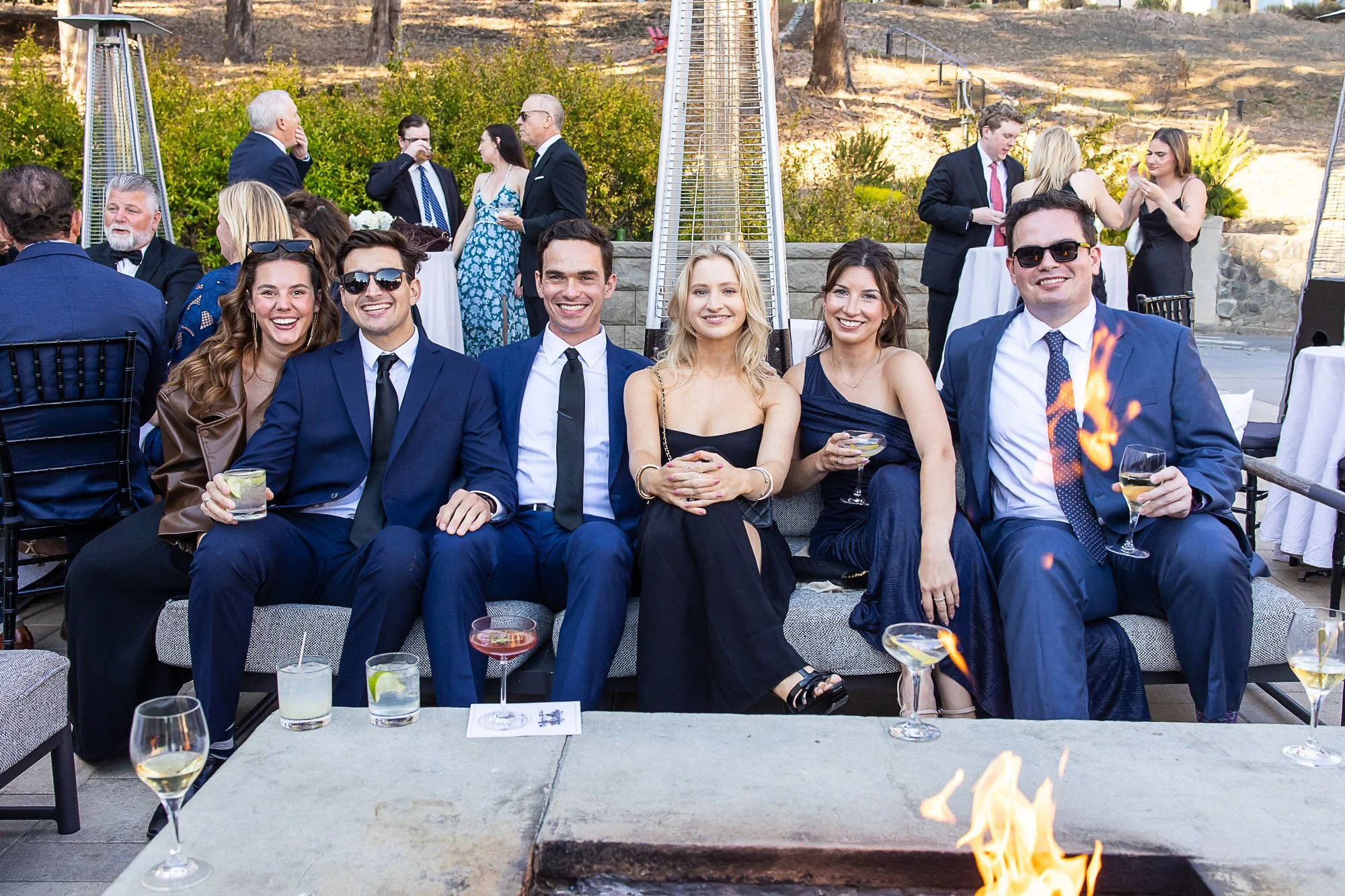 Group of young adults dressed in formal attire sitting and smiling at an outdoor social event during the evening. Some are holding drinks, and a fire feature is visible in the foreground. Additional people are mingling and conversing in the backgroun