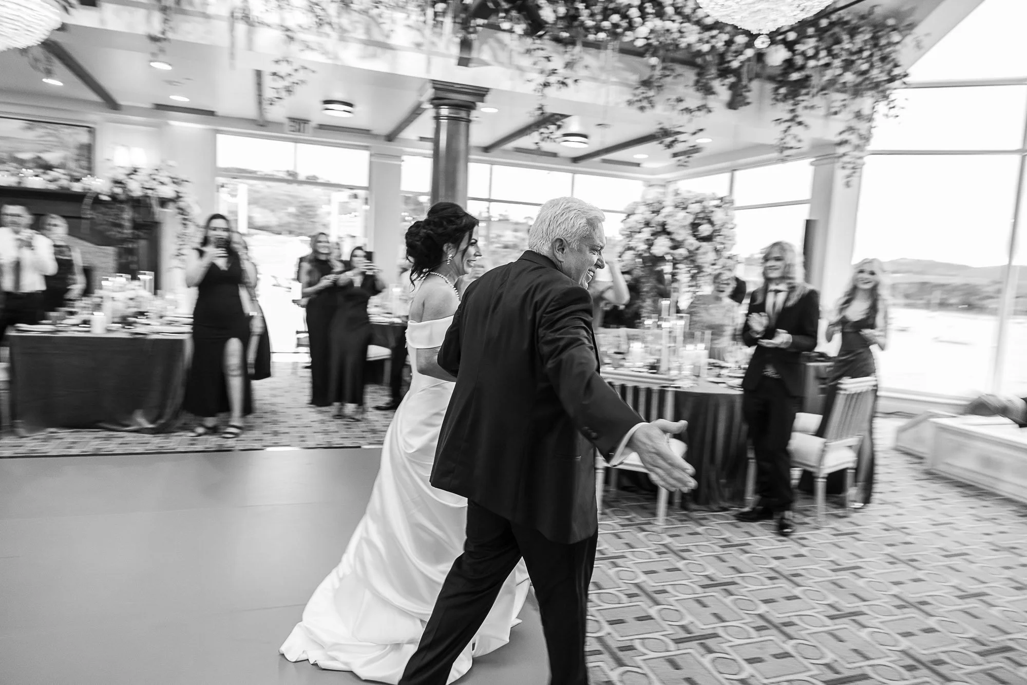 A bride and groom walk together inside the Beach and Tennis Club in Pebble Beach, CA, smiling. People are gathered in the background, some taking photos, with floral arrangements and windows letting in natural light.