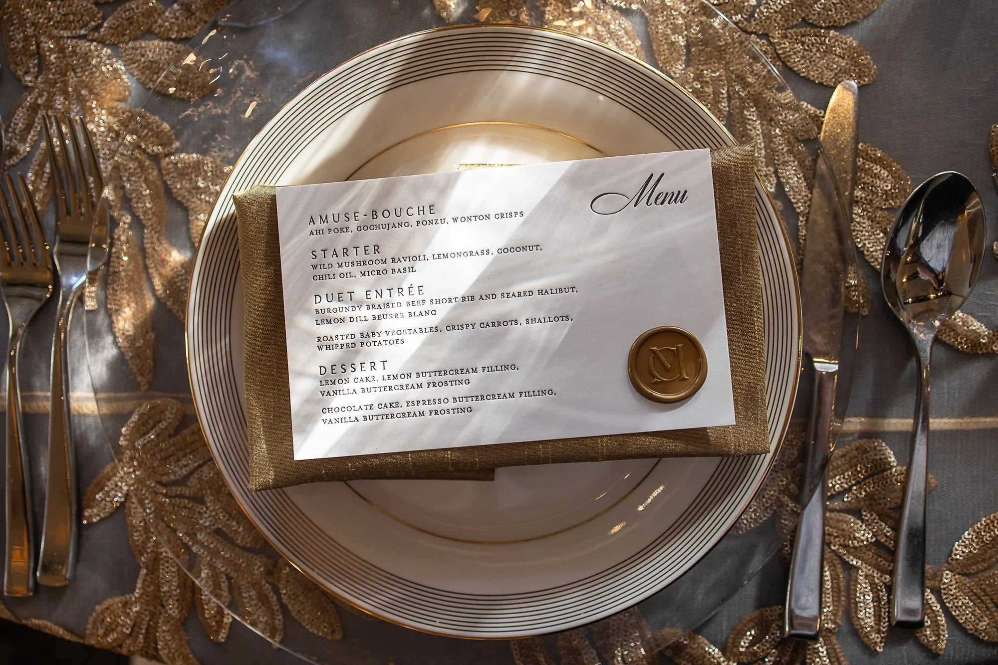 A party table setting with a white plate with black and gold stripes, a golden napkin, a menu card on top, and silverware arranged on an ornate gold and brown decorative cloth or table runner at Cavallo Point in Sausalito, CA.