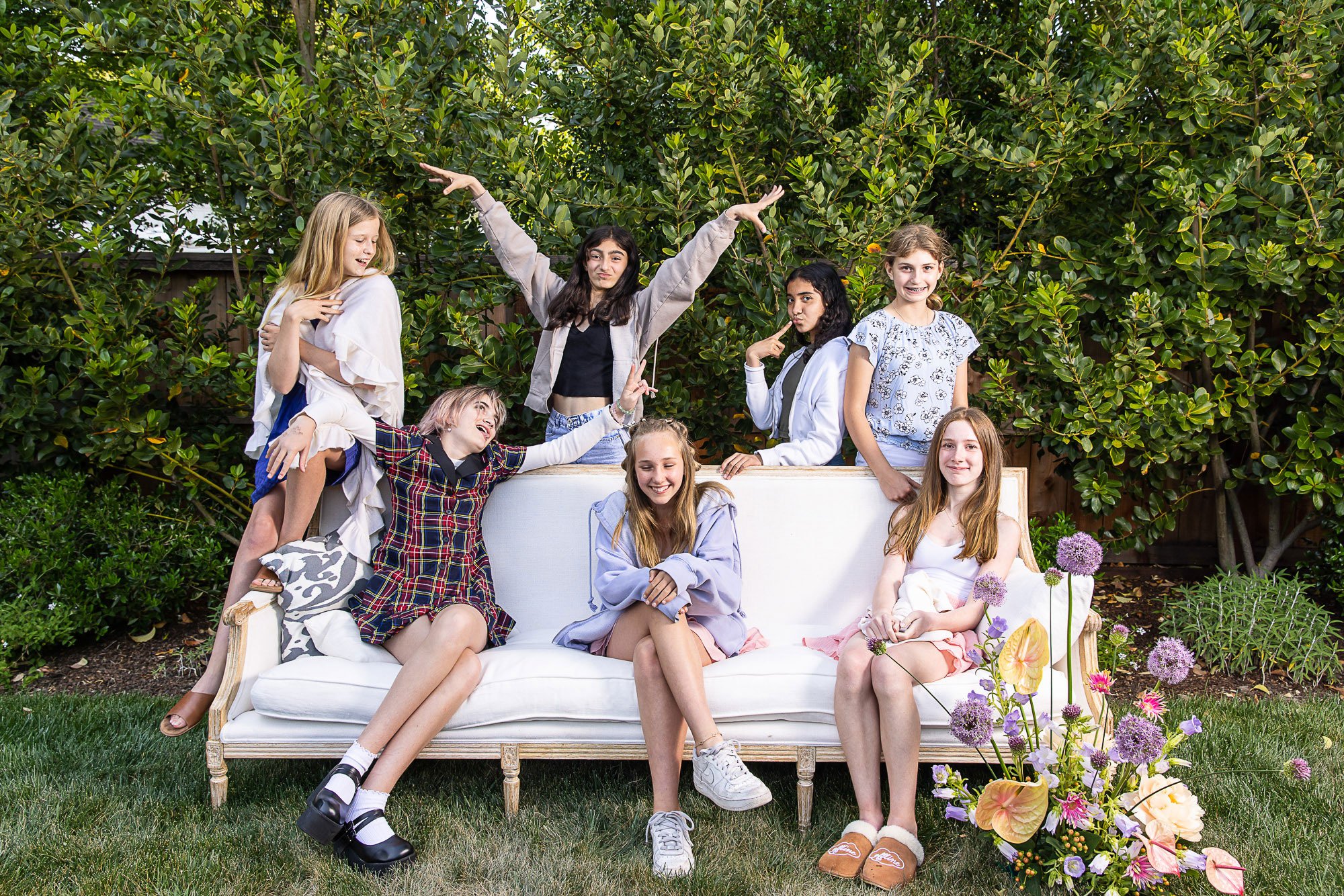 A group of seven young girls outdoors posing on and around a white vintage-style sofa, with lush green bushes and flowering plants in the background.