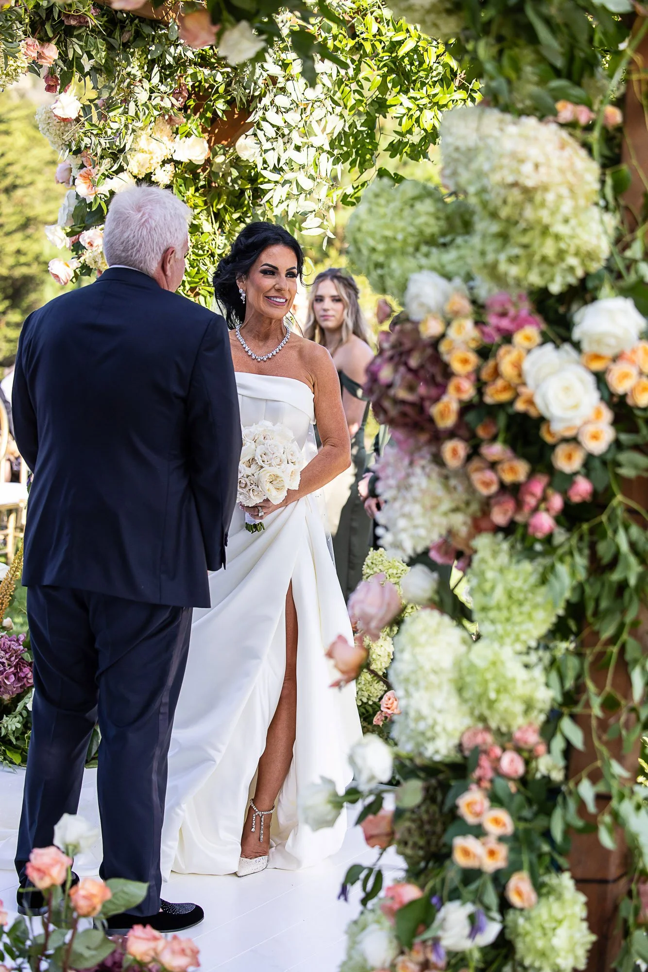 A bride wearing a white strapless wedding gown with a slit, and holding a bouquet of white roses, is smiling and standing beneath a floral arch during a wedding ceremony at the Beach and Tennis Club in Pebble Beach.