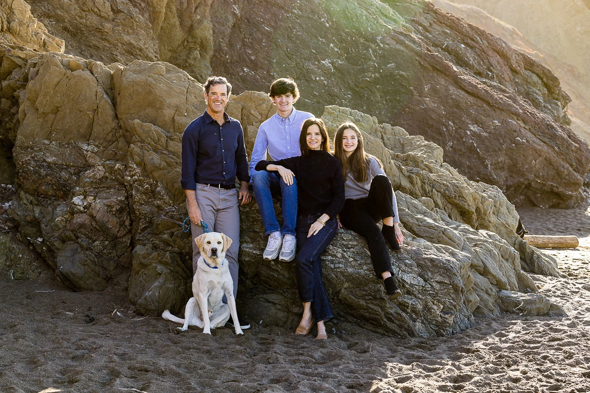 A family of four adults and one young girl with a dog sitting on rocks at the beach, with a rocky hillside in the background, during the late afternoon at Rodeo Beach in Marin California.