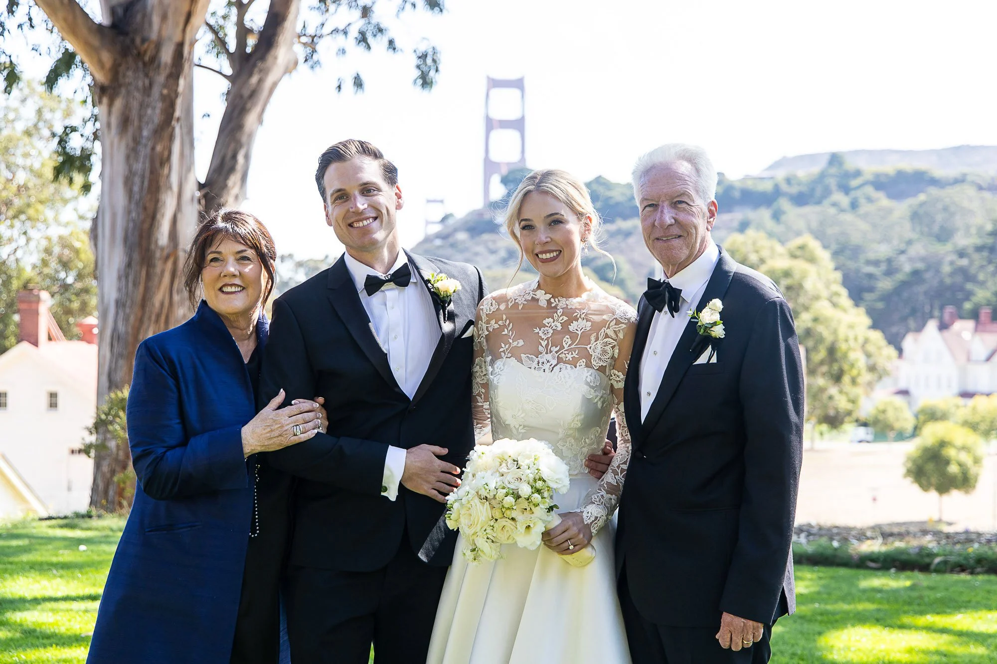 A wedding group photo featuring four adults and a bride holding a bouquet of white flowers, with the Golden Gate Bridge visible in the background on a sunny day at Cavallo Point in Sausalito, CA.