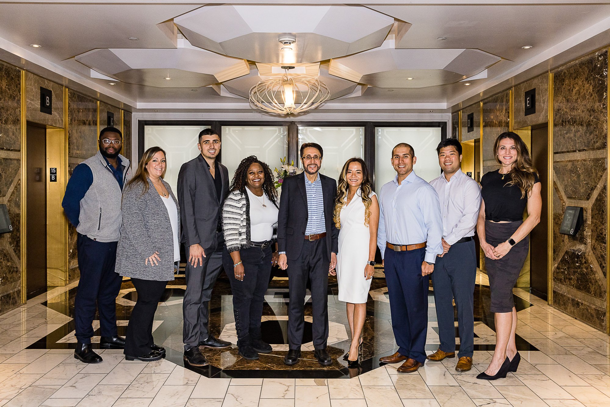 Group of nine diverse professionals standing in a hotel lobby, smiling at the camera with an elevator and marble decor behind them.