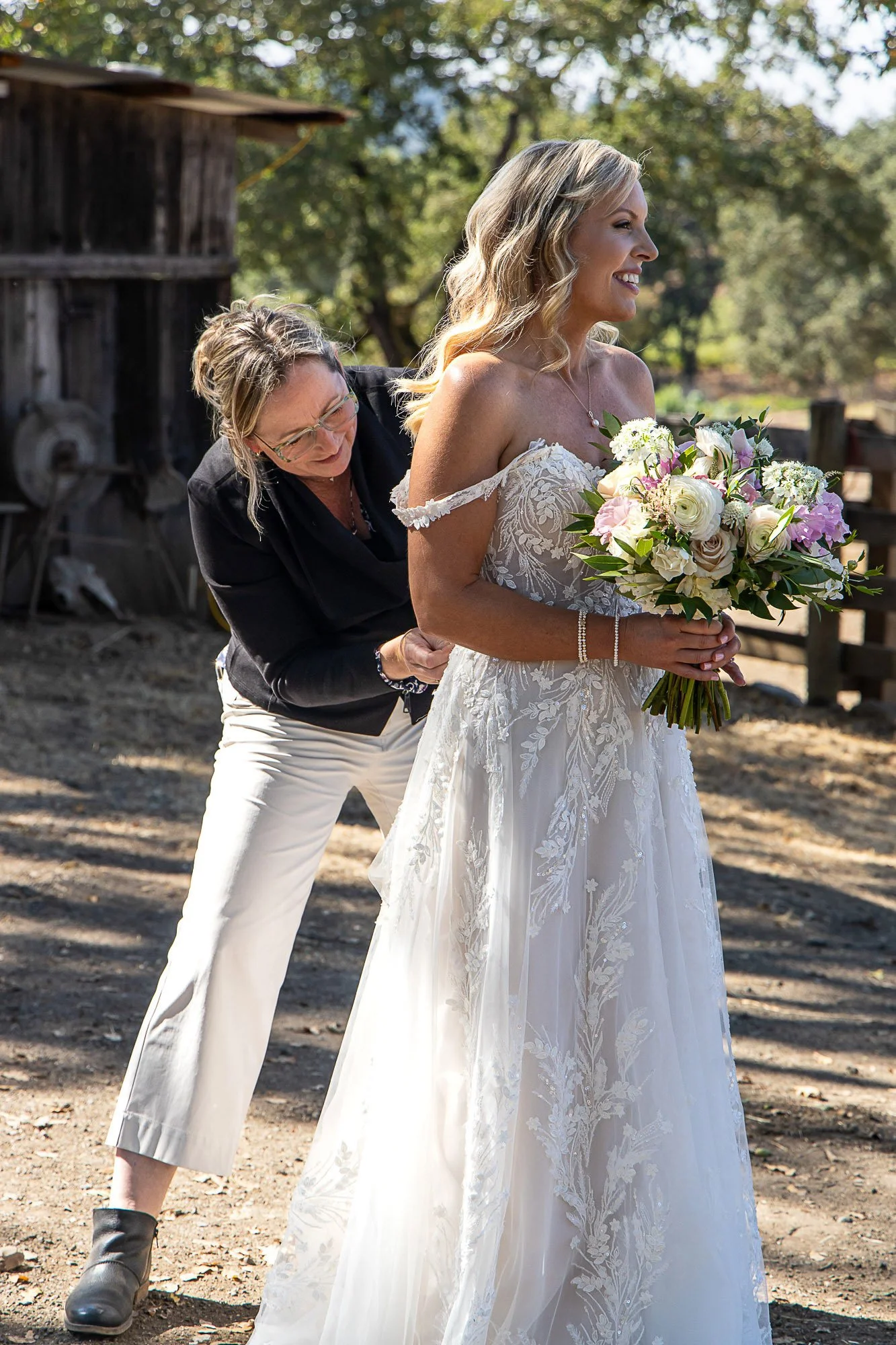 A bride in a white wedding dress holding a bouquet of flowers, standing outdoors, while Tara Arrowood in glasses and black clothing helps her with her dress.