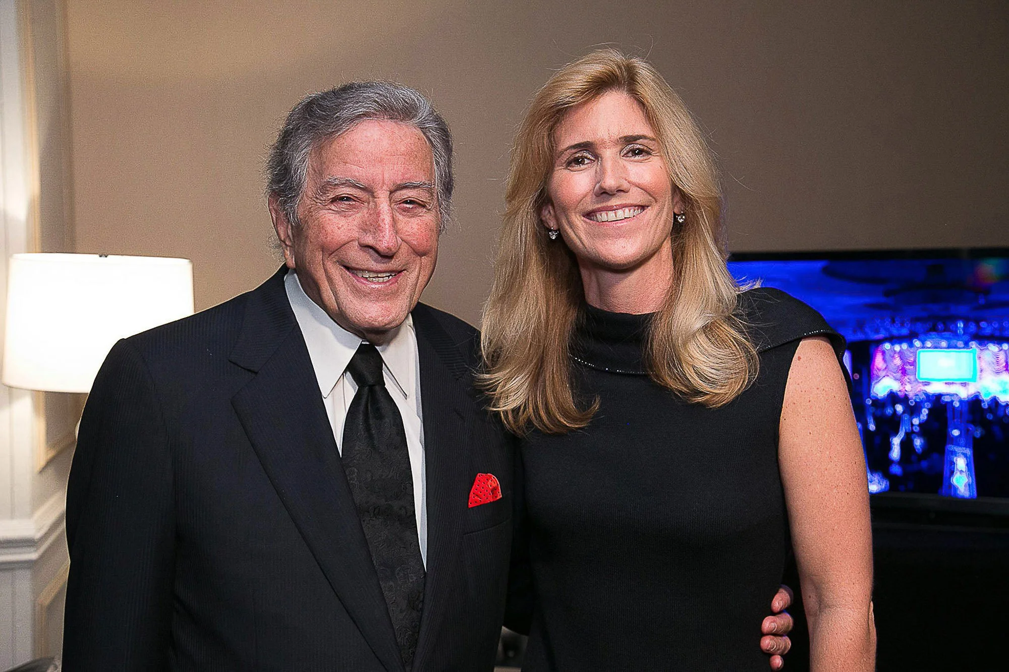 The late Tony Bennett in a suit and his wife in a black dress smiling together indoors, with a blue-lit arcade game visible in the background at the Salesforce Conference.
