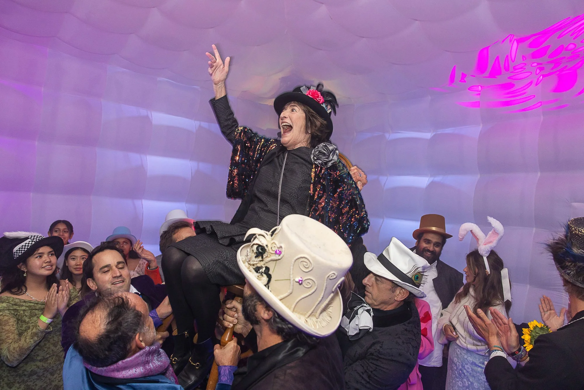 A woman in a costume and hat is being lifted by another person at a celebration, surrounded by a group of people dressed in colorful, whimsical costumes, with some wearing hats with bunny ears, at an indoor party with purple lighting.