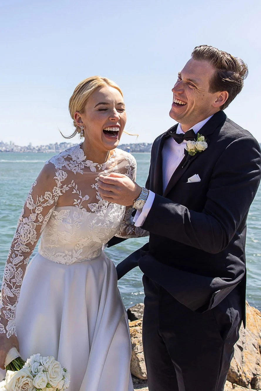 Bride laughing with her groom on the shoreline at Cavallo Point, Sausalito, with the San Francisco skyline behind them