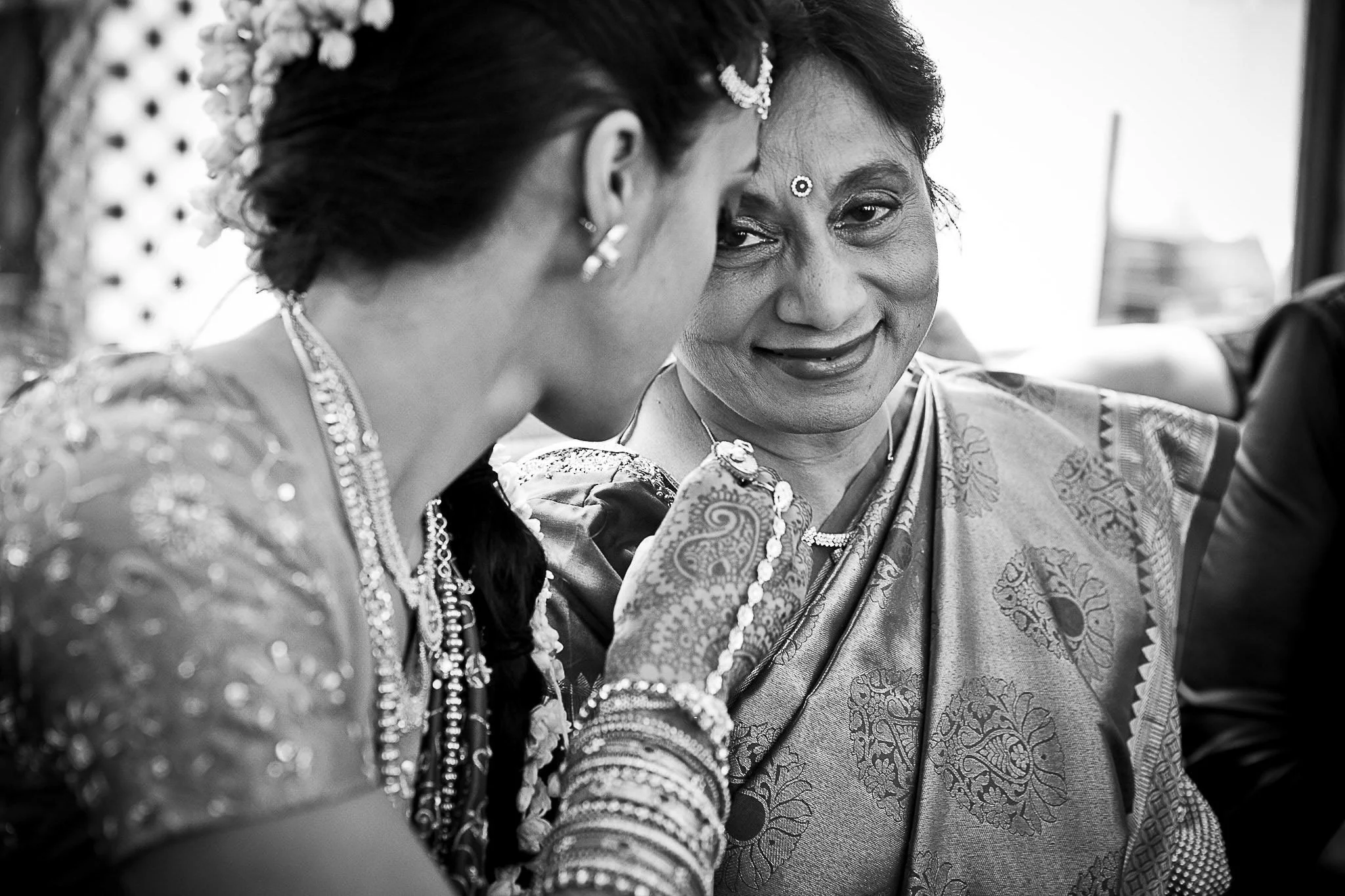 Two women dressed in traditional Indian sarees and jewelry sharing a moment at a celebration, with one woman smiling and looking at the other.