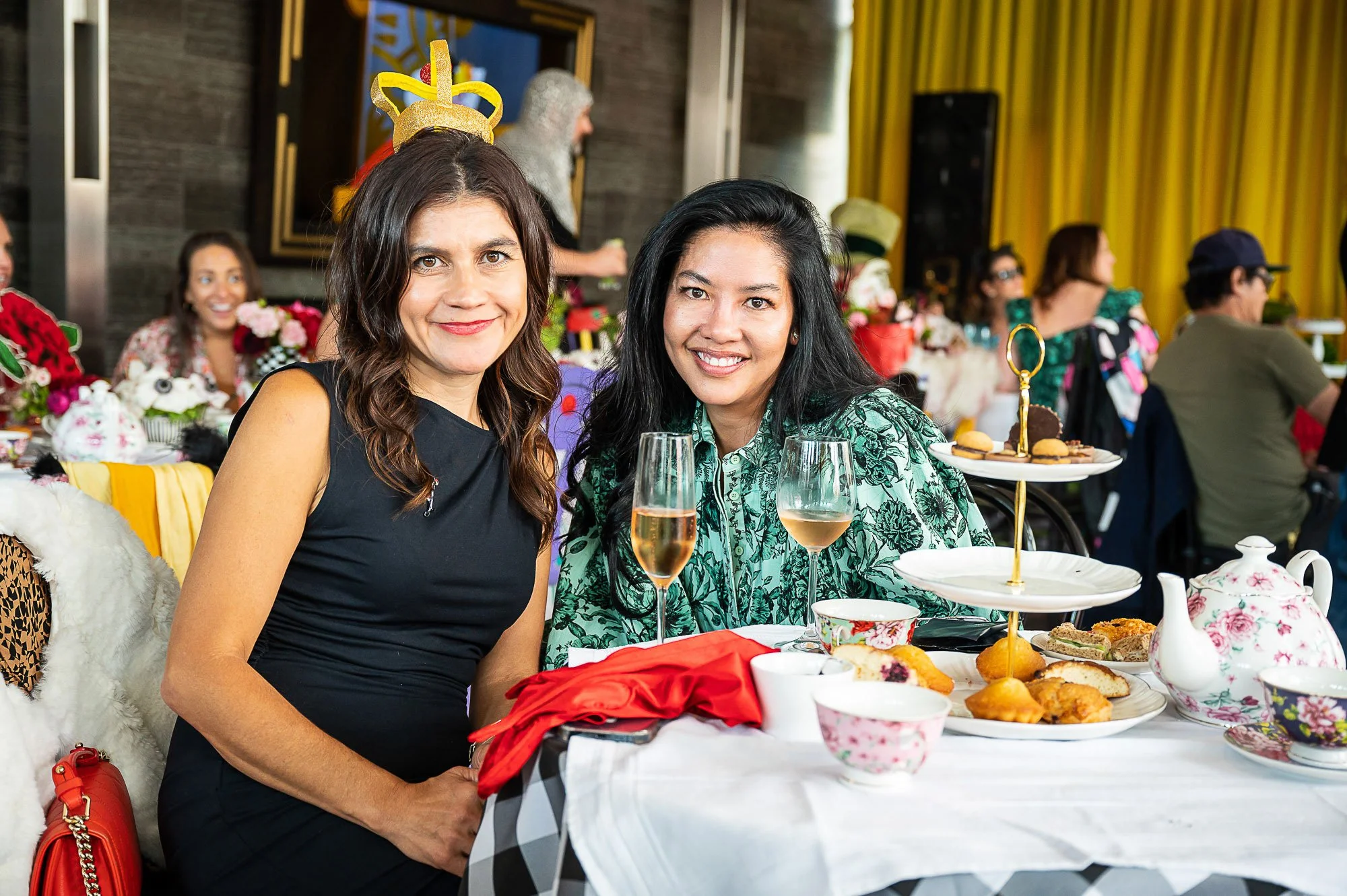 Two women sitting at a table during a celebration, smiling at the camera. They have glasses of champagne and a variety of desserts and snacks on the table. People and decorations are visible in the background.