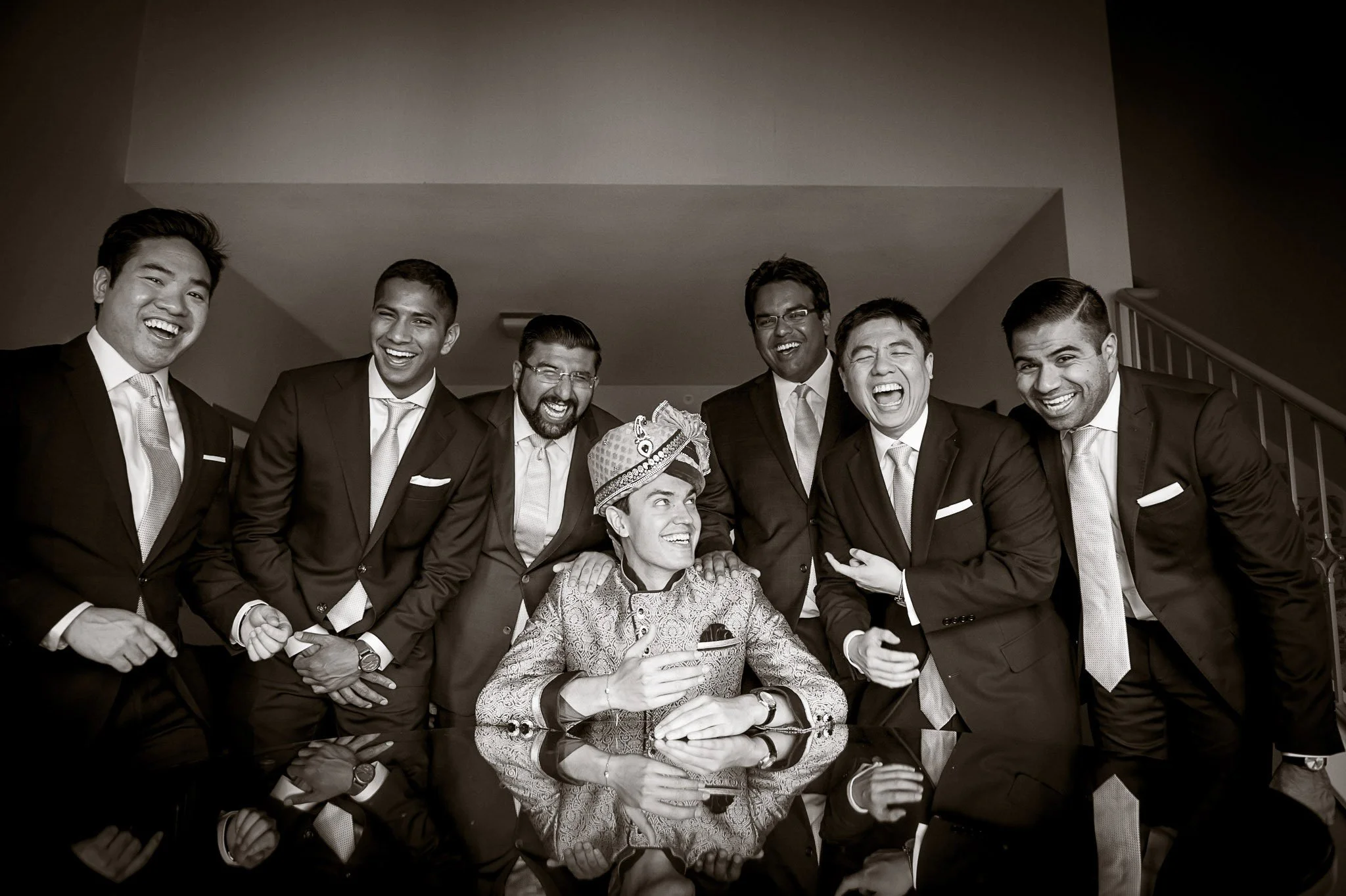A group of nine people, mostly men in suits, smiling and posing together, with one woman seated at the center wearing traditional attire and a crown, in an indoor setting.