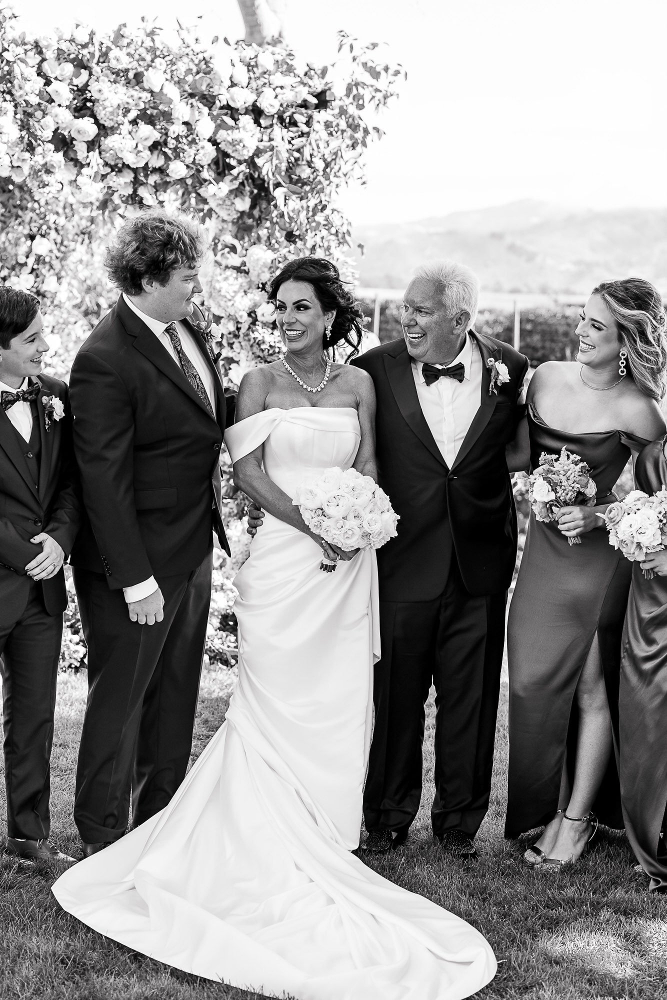 A black and white photograph of a group of people at a wedding. The bride, holding a bouquet of flowers, is smiling and looking at the groom at the Beach and Tennis Club in Pebble Beach, CA.