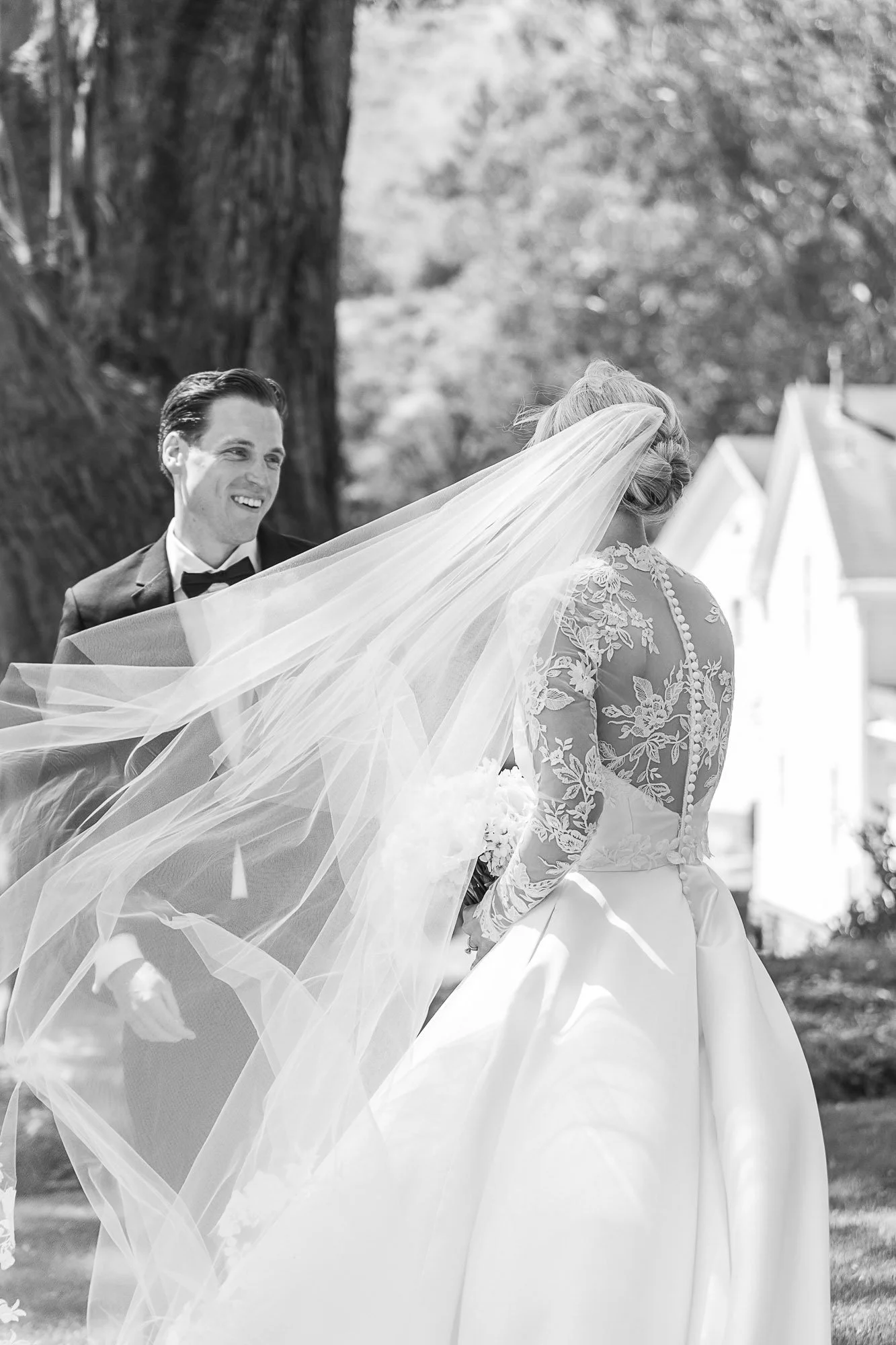A black and white photograph of a bride and groom outdoors, with the groom smiling and the bride with her back to the camera, wearing a lace gown and flowing veil at Cavallo Point in Sausalito, CA.