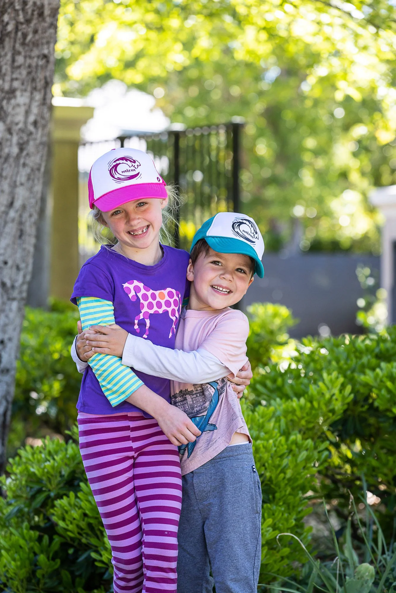 Two children, a girl and a boy, are happily hugging outdoors on a sunny day with green trees and bushes in the background. Both are wearing colorful hats and casual clothes, smiling at the camera.