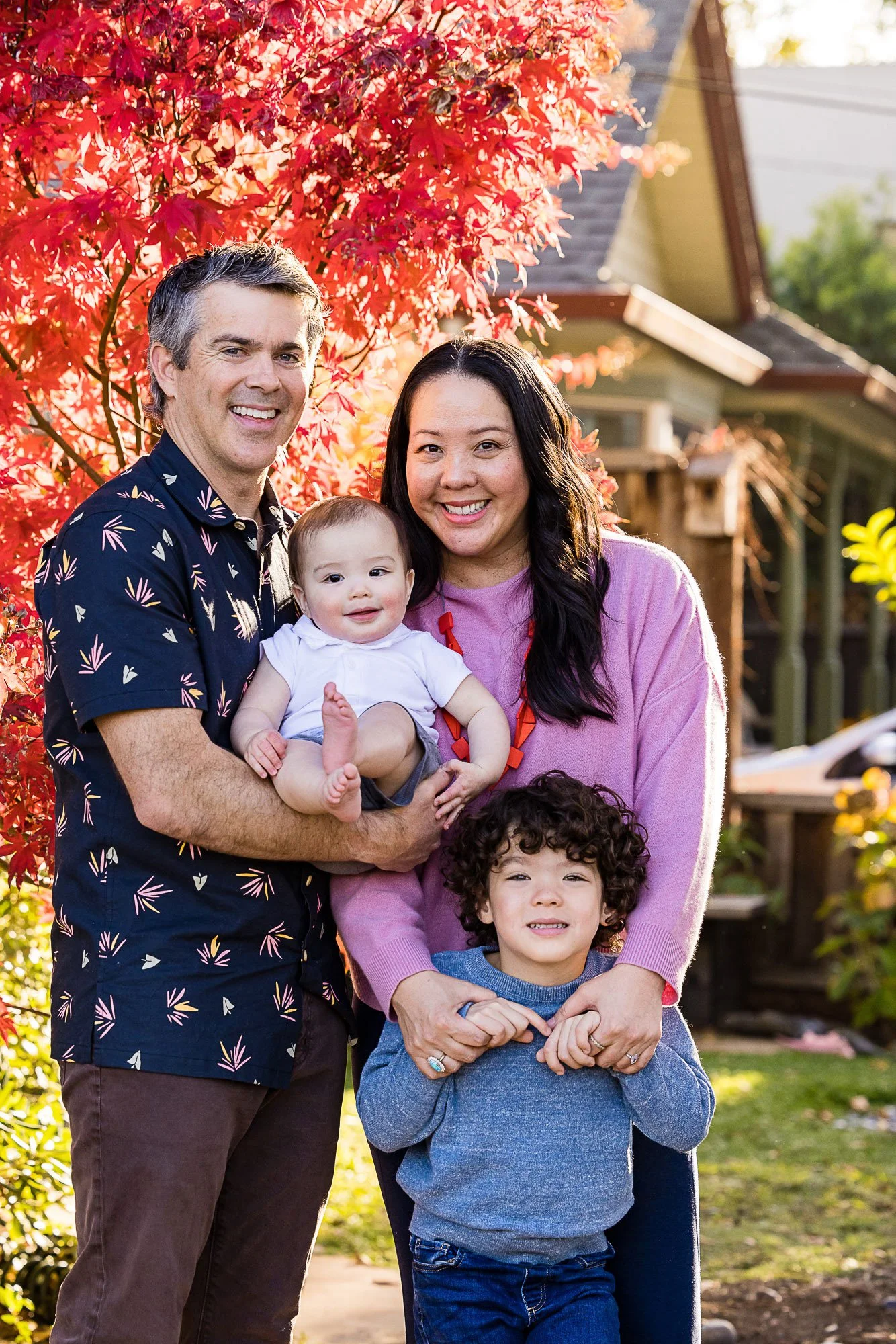 A happy multi-ethnic family of six standing outside in front of vibrant red autumn leaves, smiling at the camera.