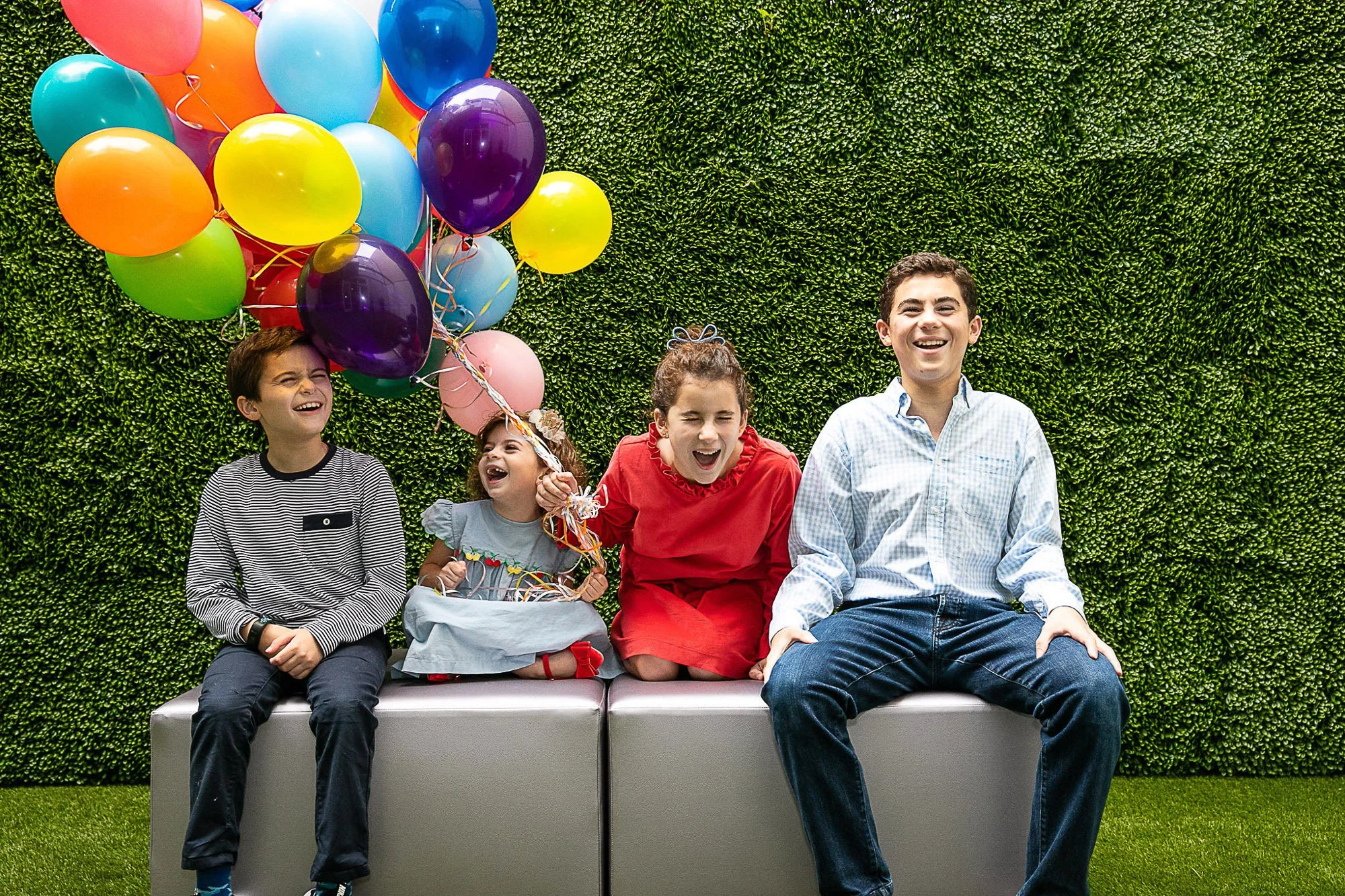 Four children and one adult sitting on a bench against a green hedge wall, with a bunch of colorful balloons held by one of the children. The children are smiling and laughing.