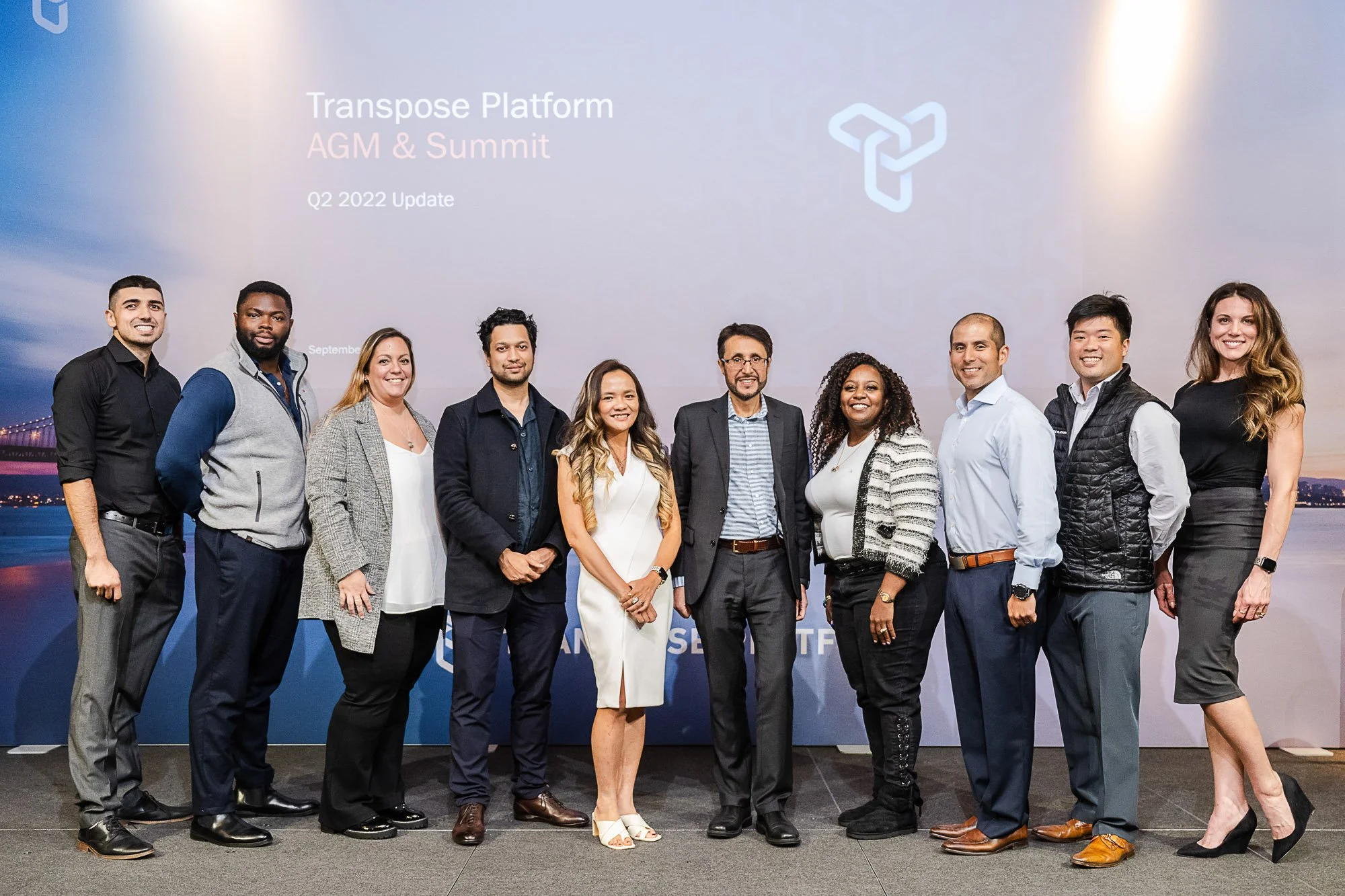 A diverse group of eleven people standing together on a stage at a professional event, with a large screen behind them displaying a company logo and the words 'Transpose Platform AGM & Summit' and 'Q2 2022 Update'.