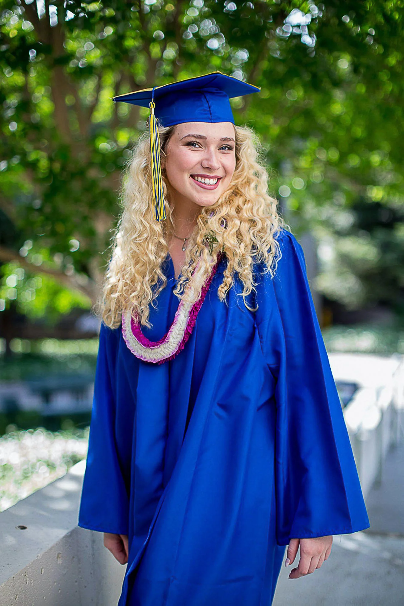 A young woman with long, curly blonde hair wearing a blue graduation cap and gown, standing outdoors with green trees in the background, smiling at the camera.