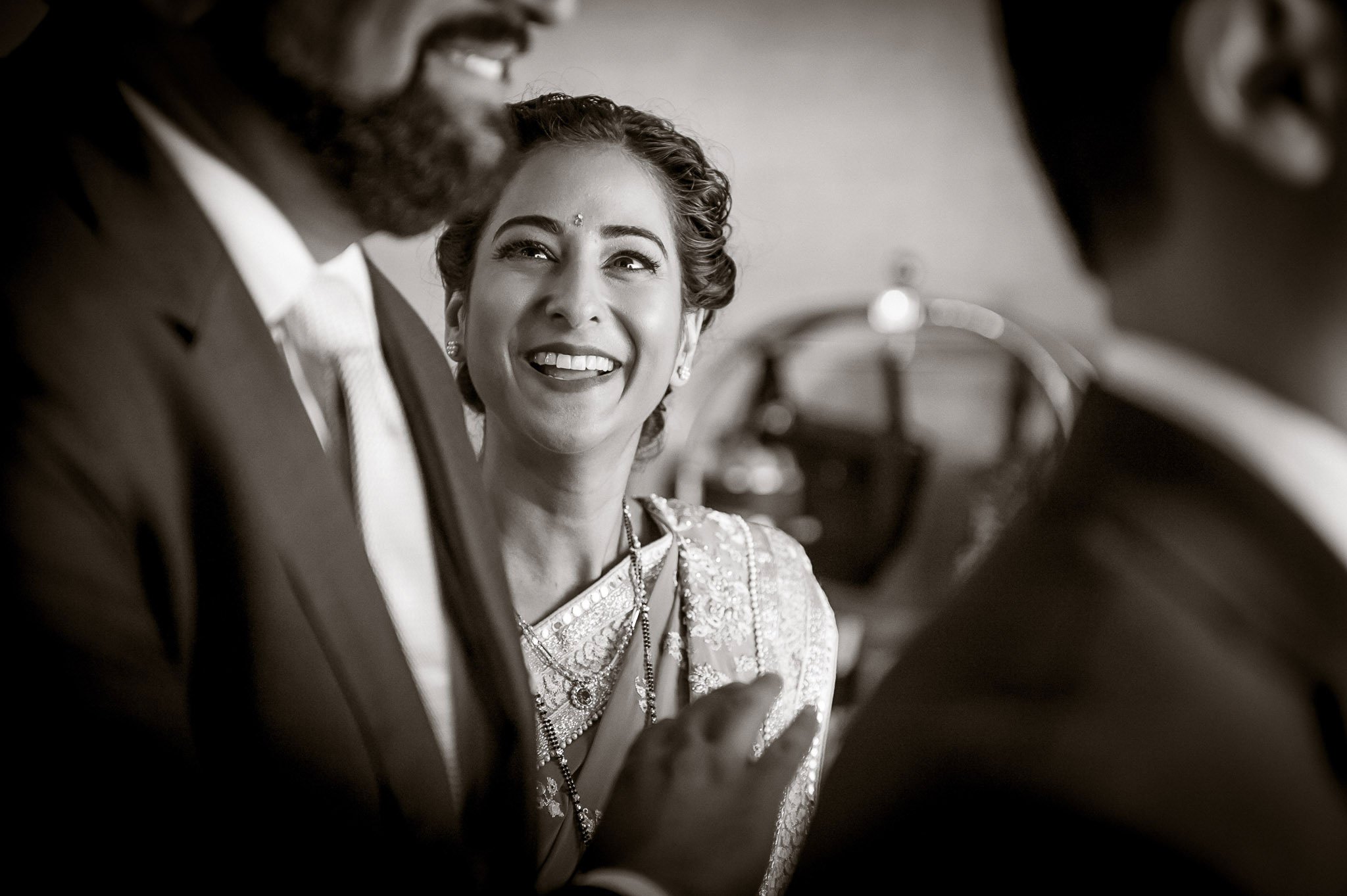 Black and white close-up photo of a smiling woman in traditional Indian attire, with her hair styled in an elegant updo, and wearing jewelry, engaging in conversation with a man in a formal suit beside her.
