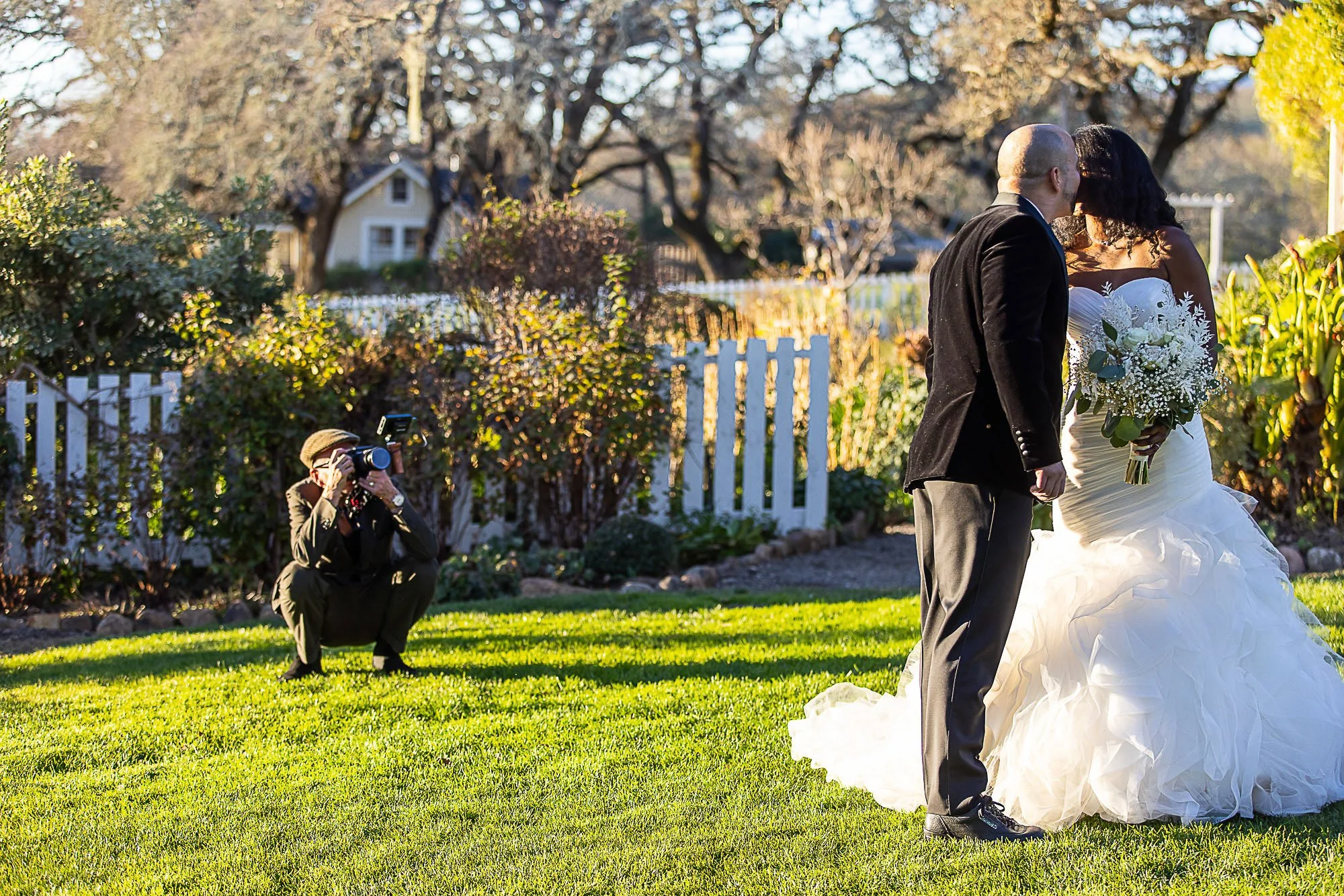A couple getting married outdoors in a garden on a sunny day. The woman is in a white wedding dress holding a bouquet, and the man is in a black suit. Videographer Martin is kneeling on the grass taking pictures.