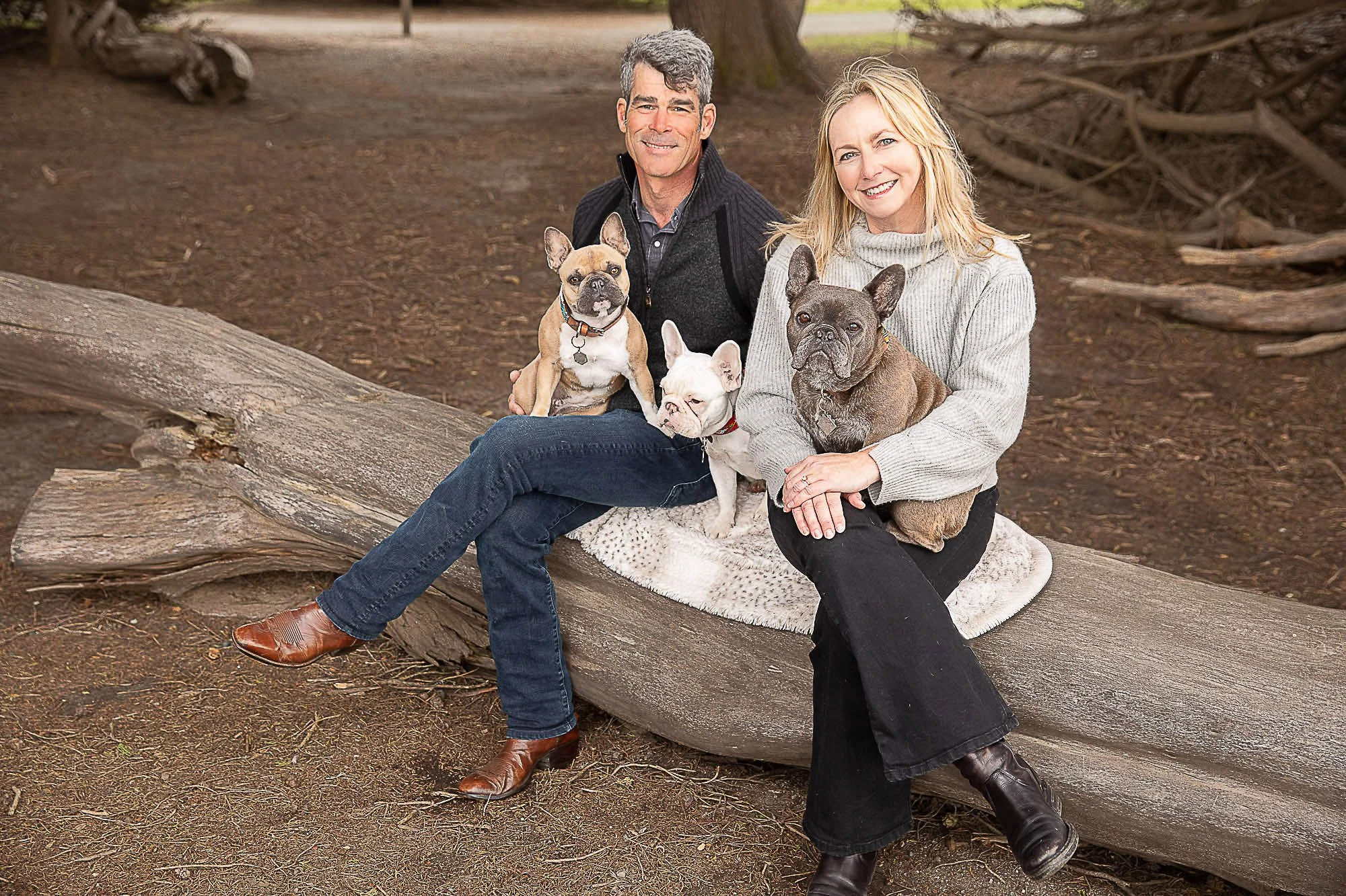 Travis Pynn and Tara Arrowood sitting on a large fallen tree trunk in a park, each holding a French Bulldog, with a natural park background of trees and dirt ground.