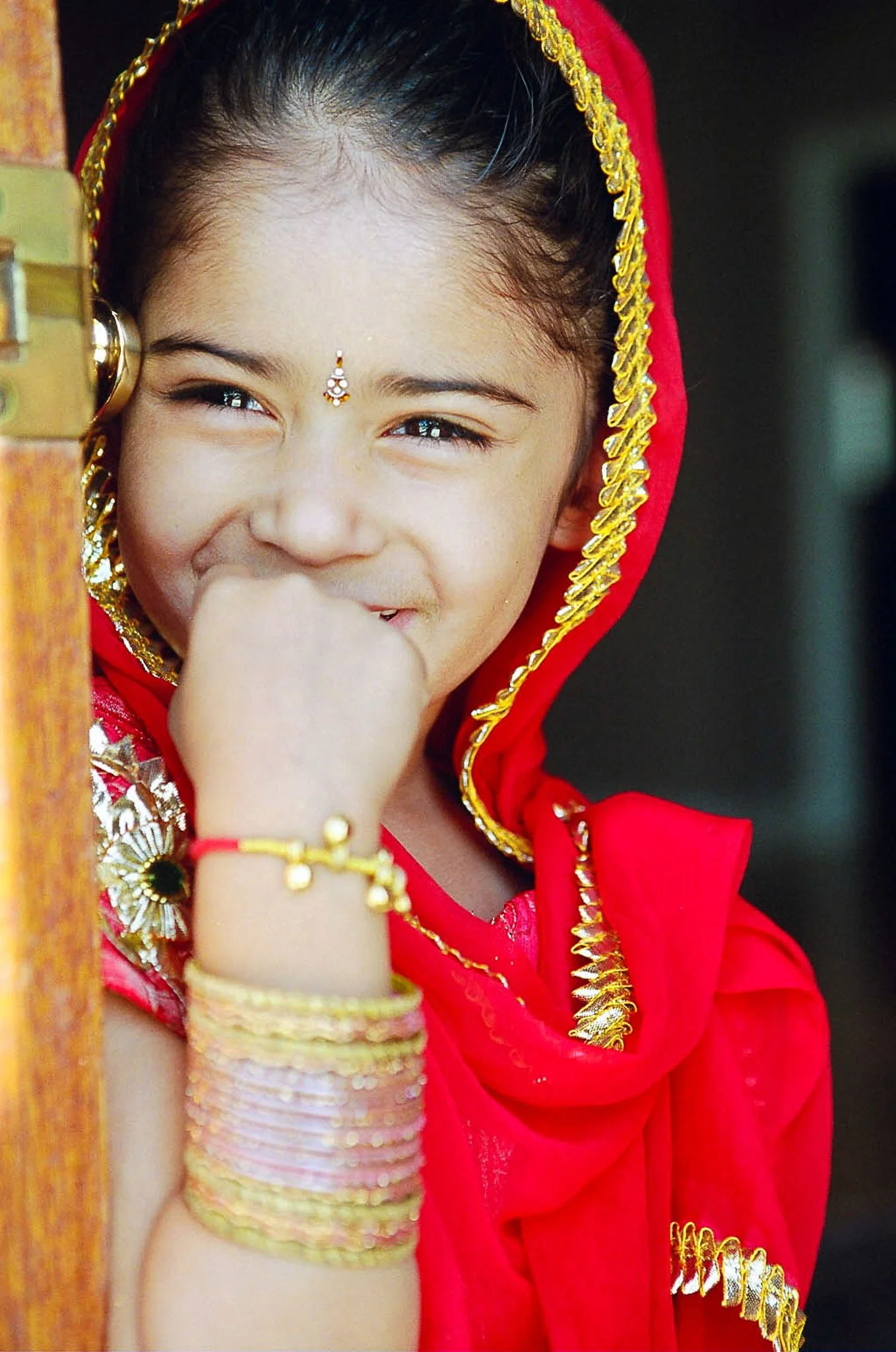 Close-up of a smiling young girl with a small traditional bindi on her forehead, wearing a red outfit with gold embellishments, and accessorized with gold jewelry, including bangles and earrings.