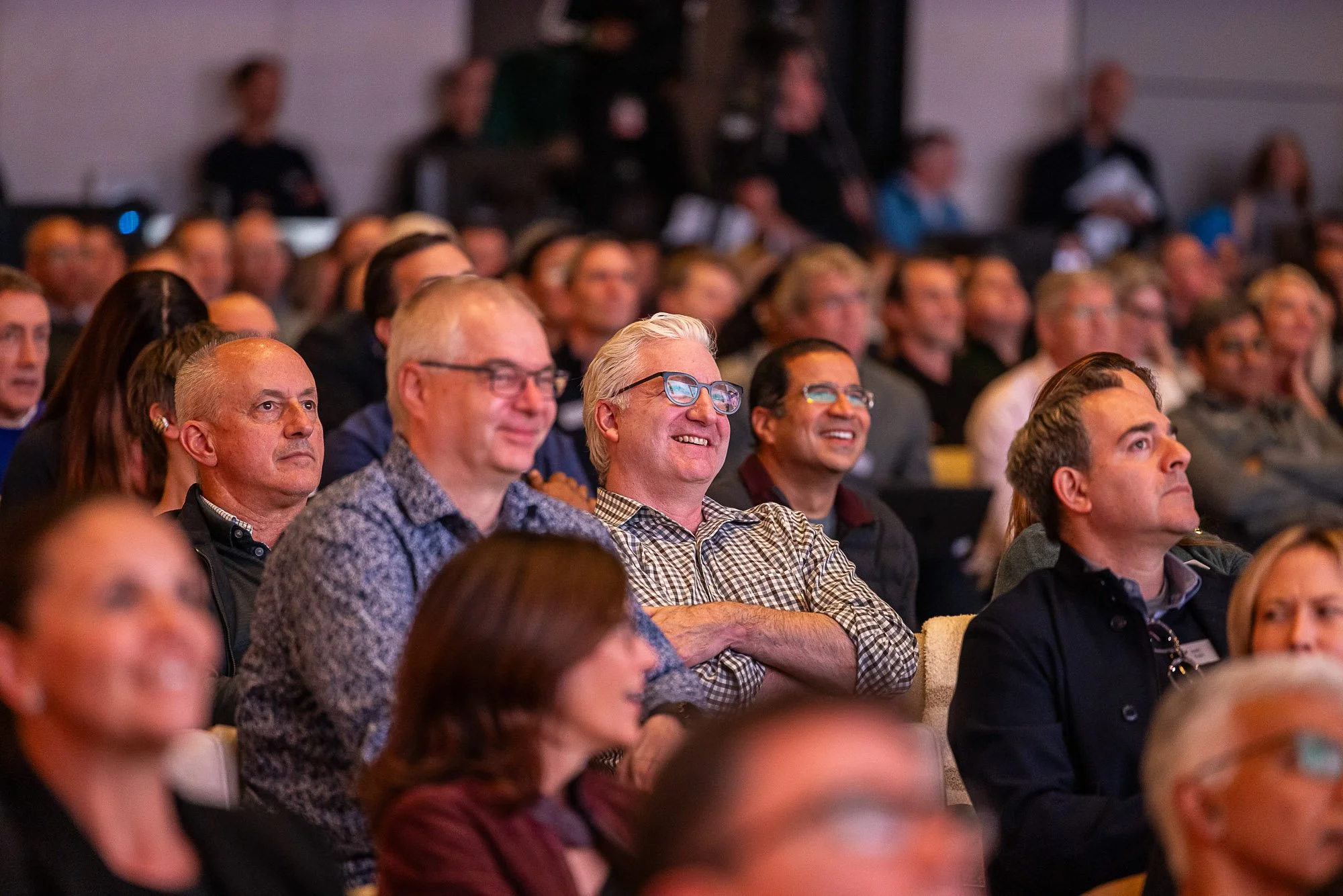 Audience members attending a conference or seminar, seated and facing forward, with some smiling and others attentively listening.