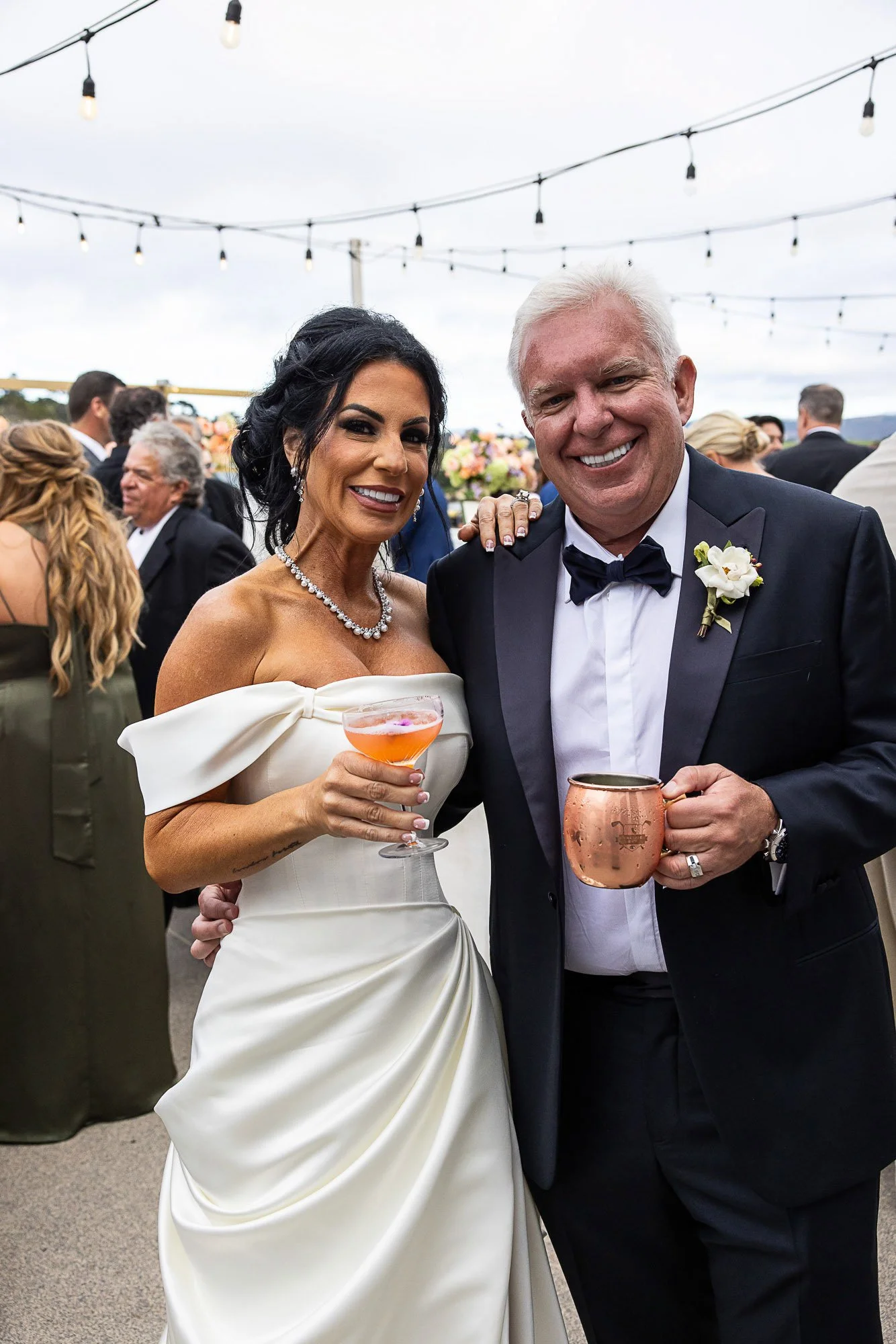 A bride and groom at an outdoor wedding reception at the Beach and Tennis Club in Pebble Beach, smiling and holding drinks, with guests in the background and string lights overhead.
