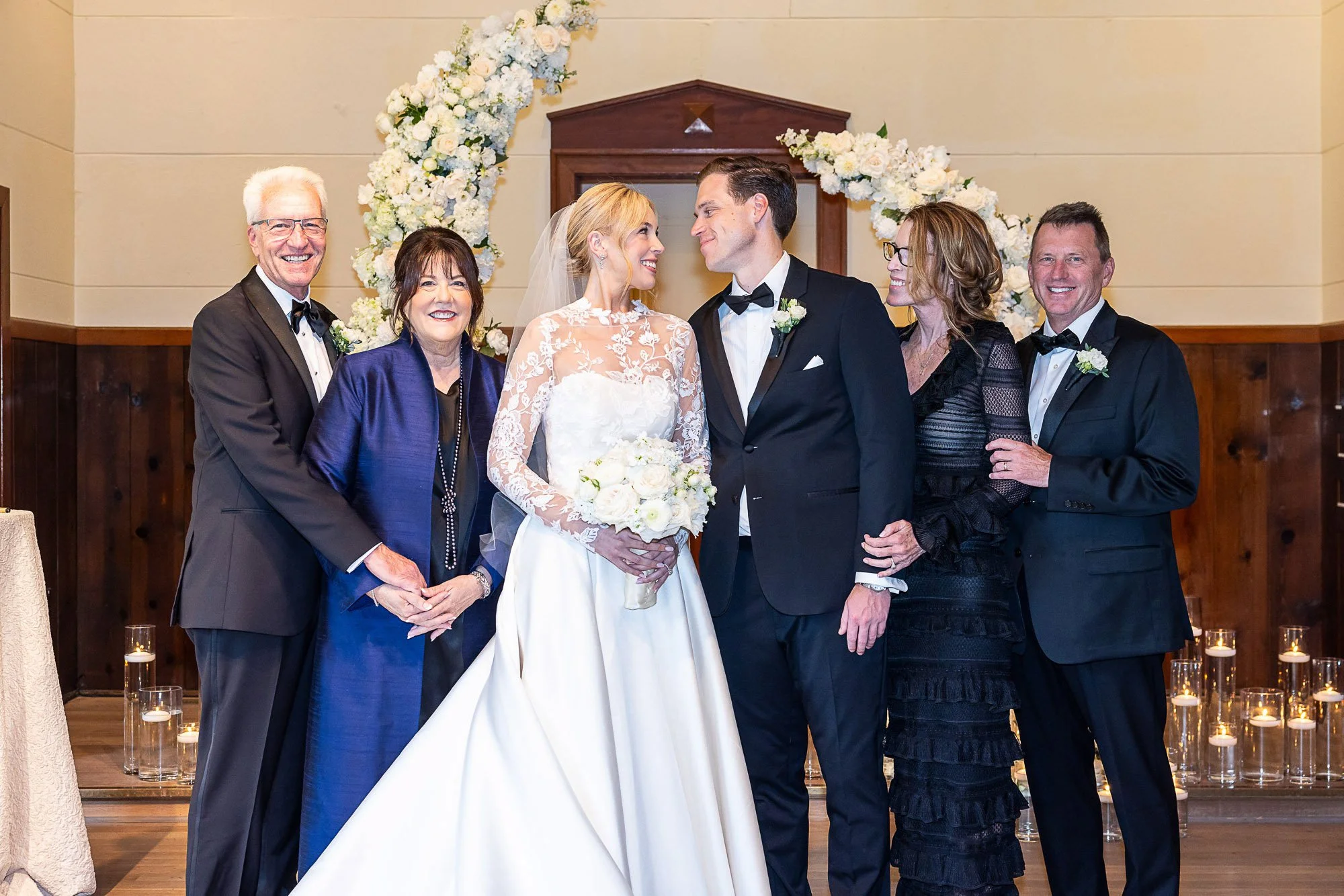 A group of seven people at a wedding, with the bride and groom in the center, surrounded by family members, in front of a floral arch and candles at Cavallo Point in Sausalito, CA.