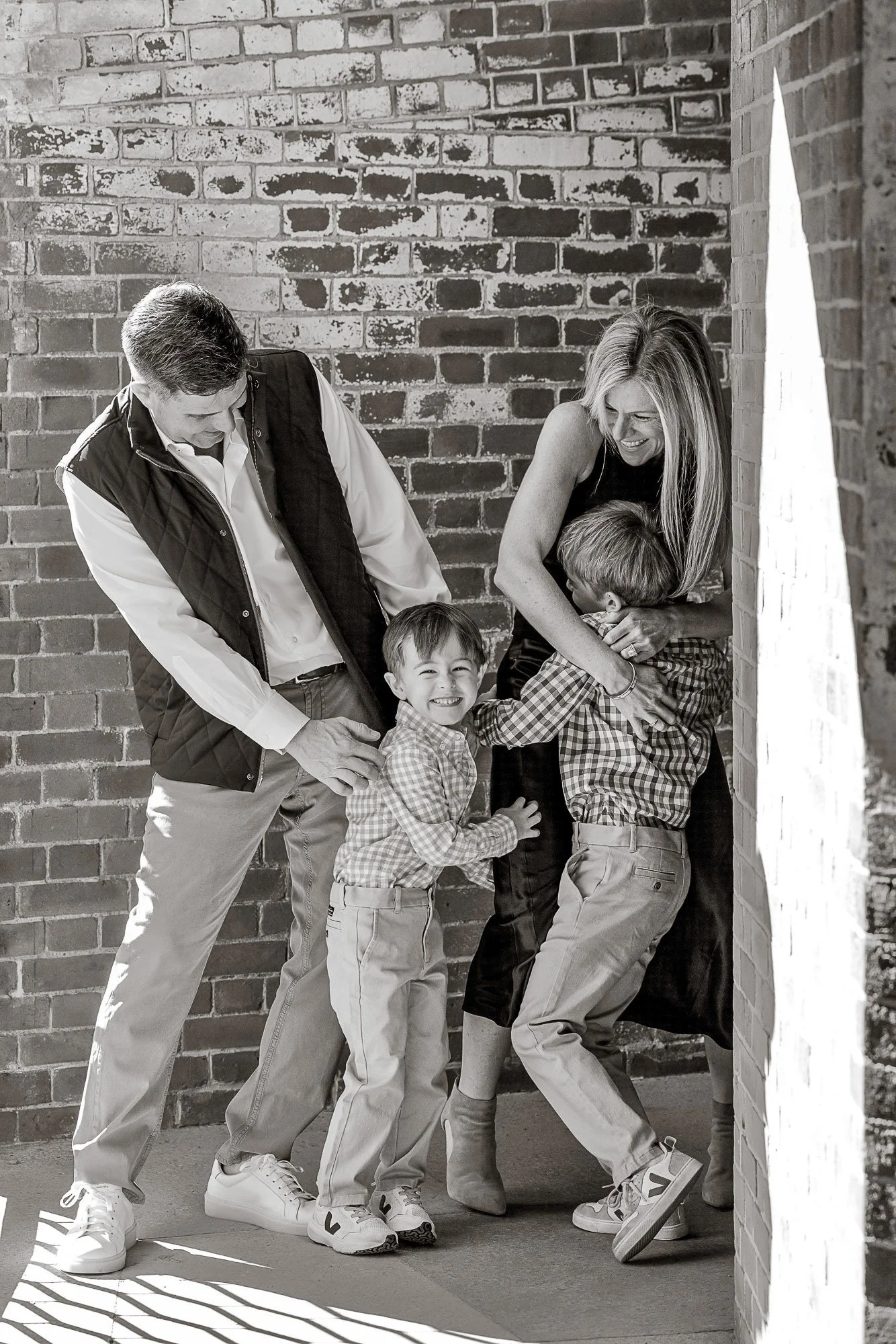A happy family of four, including two young boys, poses and interacts playfully against a brick wall, with shadows cast on the ground by sunlight.