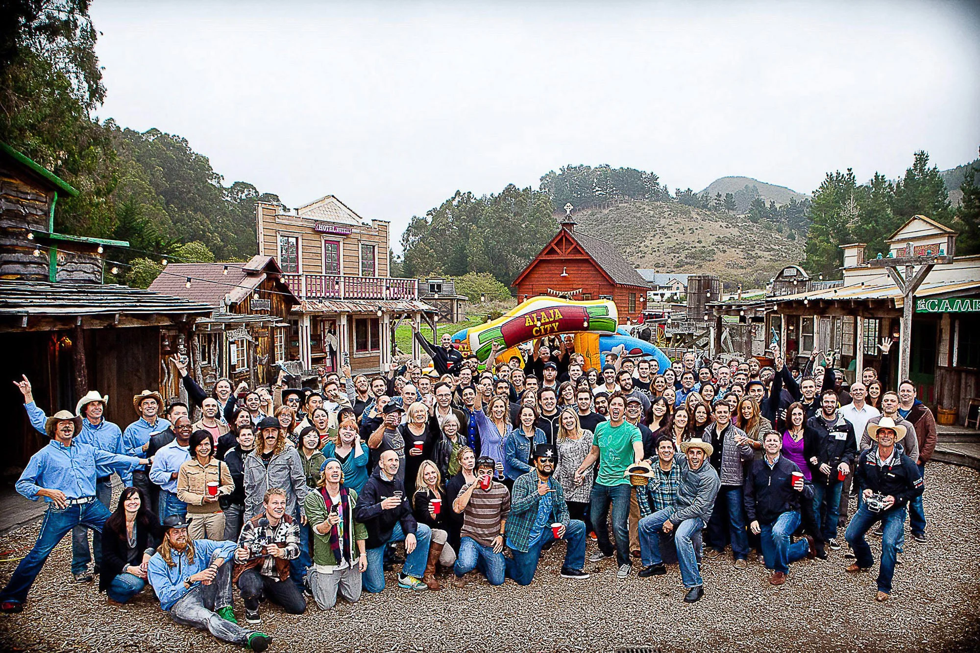 A large group of diverse people gathered outdoors in a western-themed town, smiling and posing for a photo. They are standing on a gravel street lined with wooden buildings, surrounded by hills and trees at Long Branch Farms in Half Moon Bay.