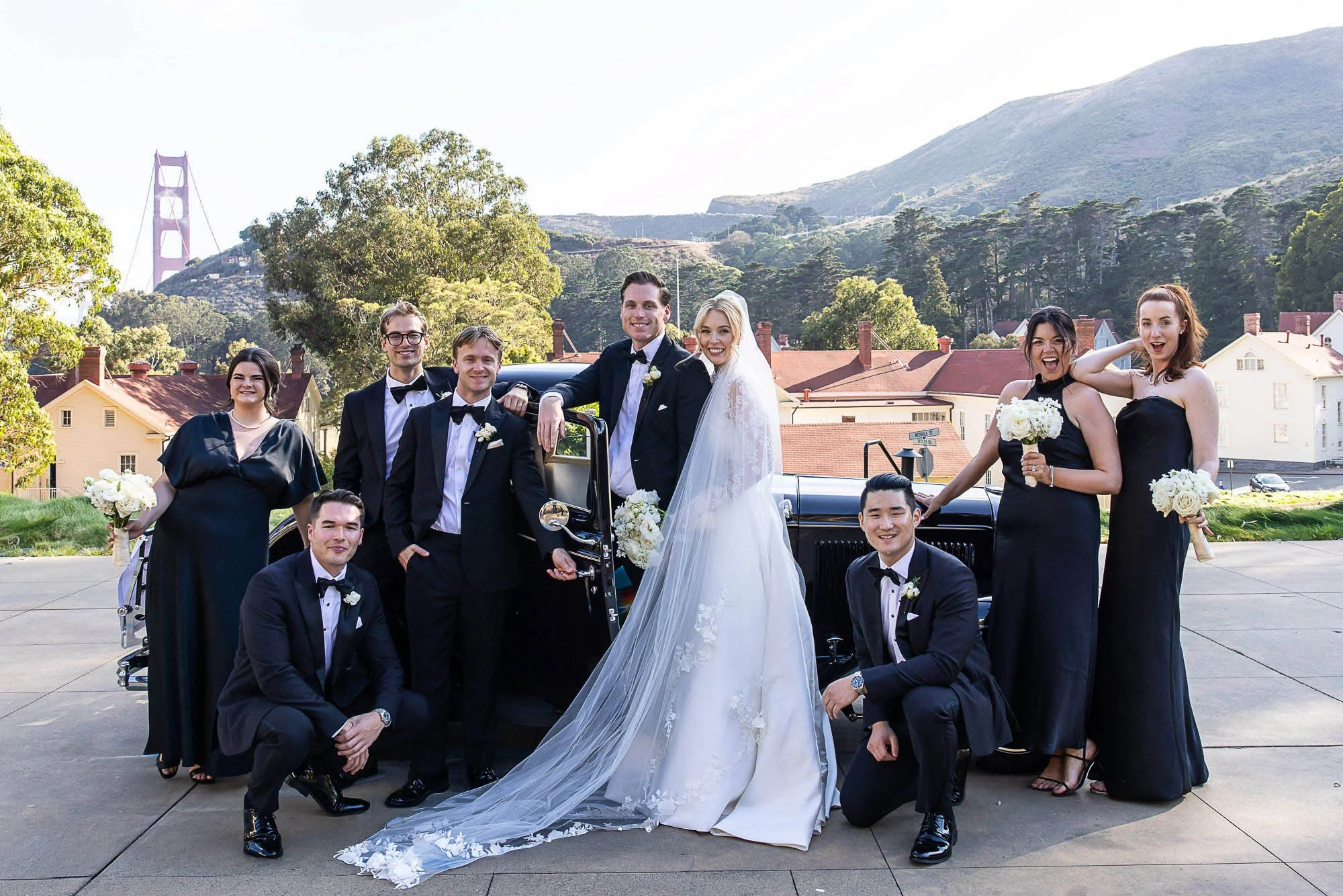 A bride and groom with wedding party in front of a classic car at Cavallo Point in Sausalito, with a mountain and the Golden Gate Bridge in the background.