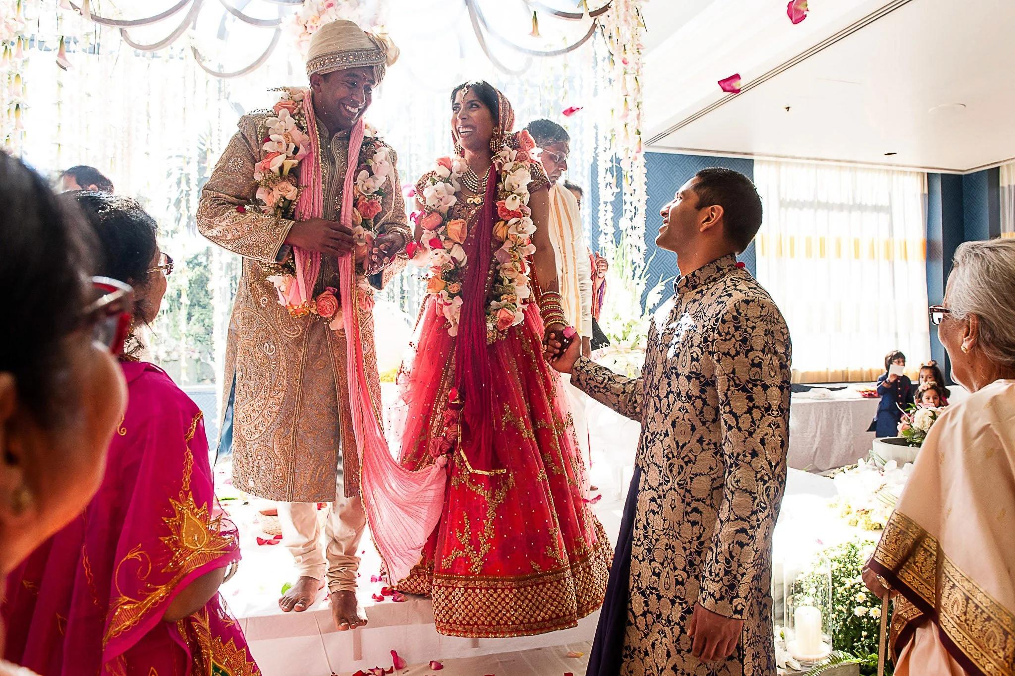 A bride and groom in traditional Indian wedding attire exchanging flower garlands during a wedding ceremony, surrounded by family and friends.