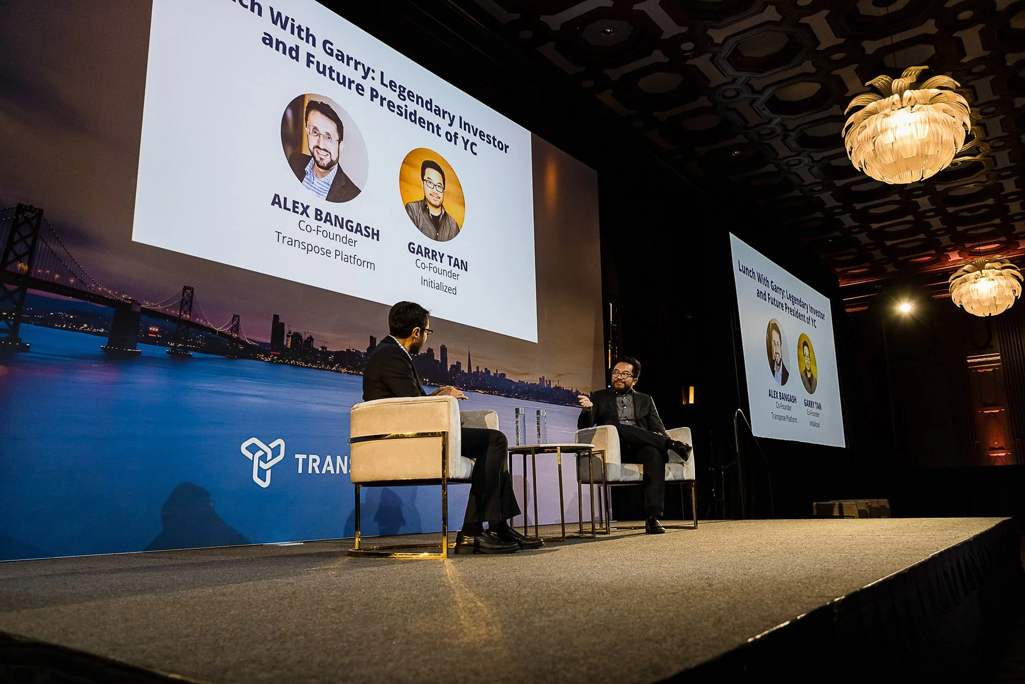 Two men sitting on stage in discussion at a conference, with large screens behind them displaying profiles of Alex Bangash and Garry Tan, and a cityscape background with a bridge, at a formal event under ornate ceiling lighting.