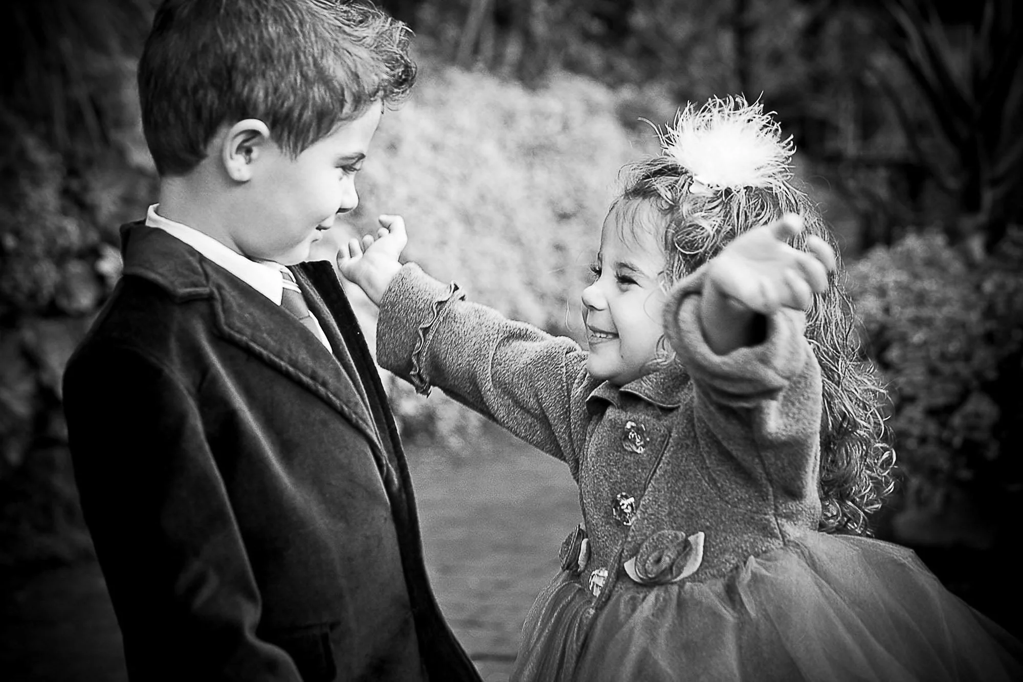 A black-and-white photo of a boy and a girl smiling and facing each other outdoors. The girl is reaching out her hand and touching the boy's chest with her fingertips. The boy is looking at her with a smile. The girl has curly hair tied with a big fl