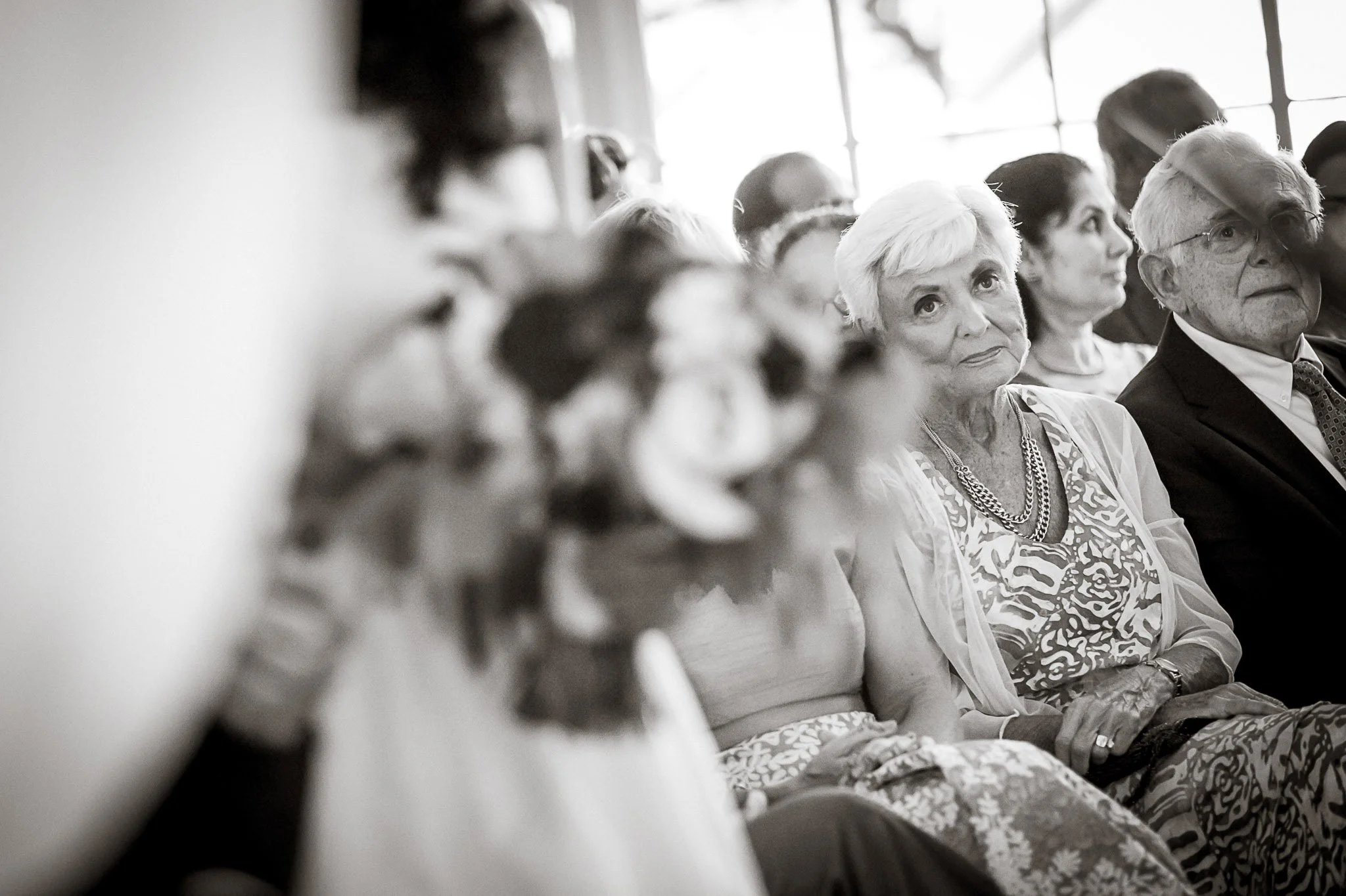 Black and white photo of elderly women and men sitting in a row, attending a formal event or gathering, with a window in the background.