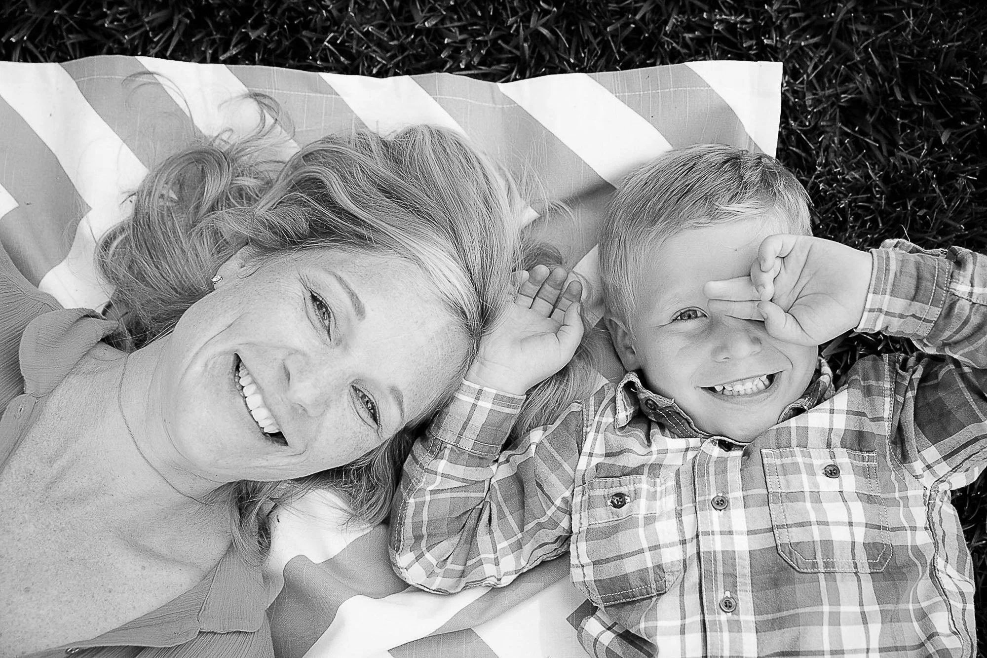 A smiling woman with wavy hair lying on a striped blanket outdoors with a young boy, both making playful gestures with their hands.
